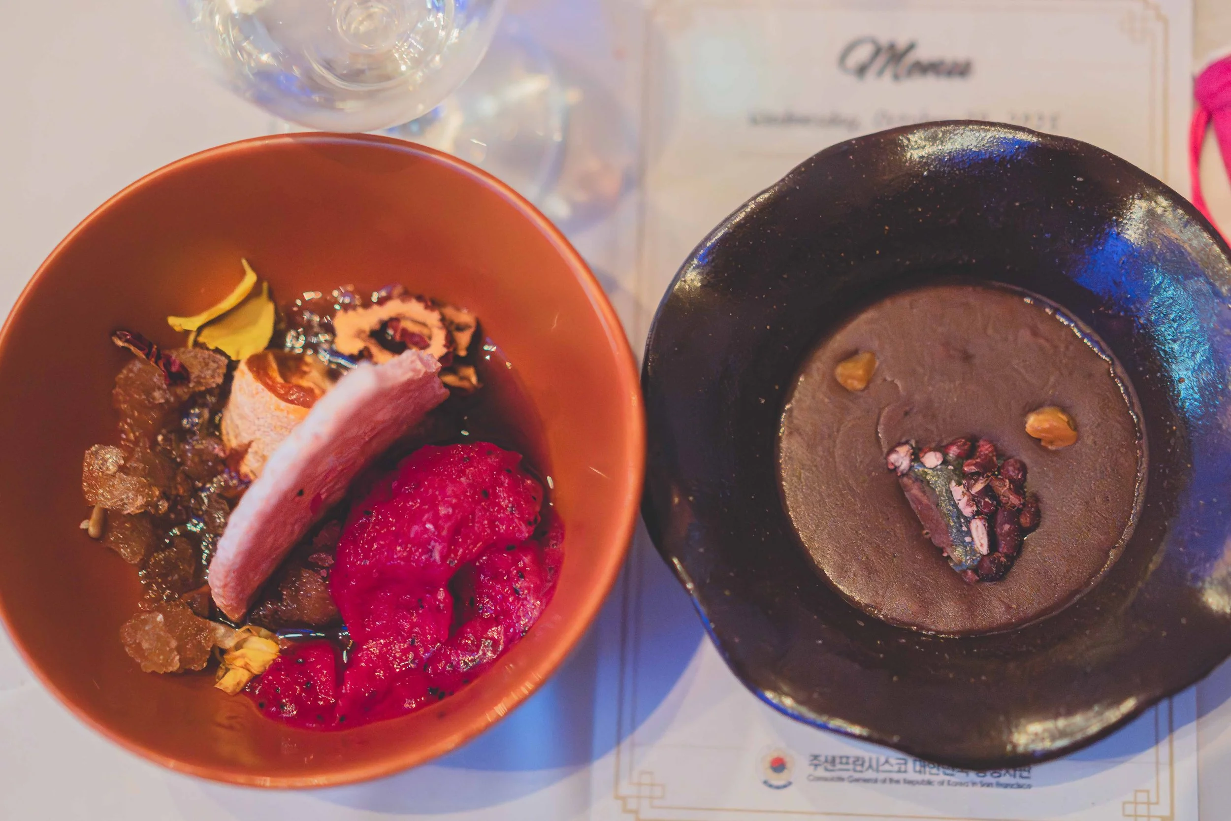 Two bowls of Japanese dessert, one black with chocolate and nuts, one orange with assorted fruit and sweets, on a white table with a glass of water in the background.