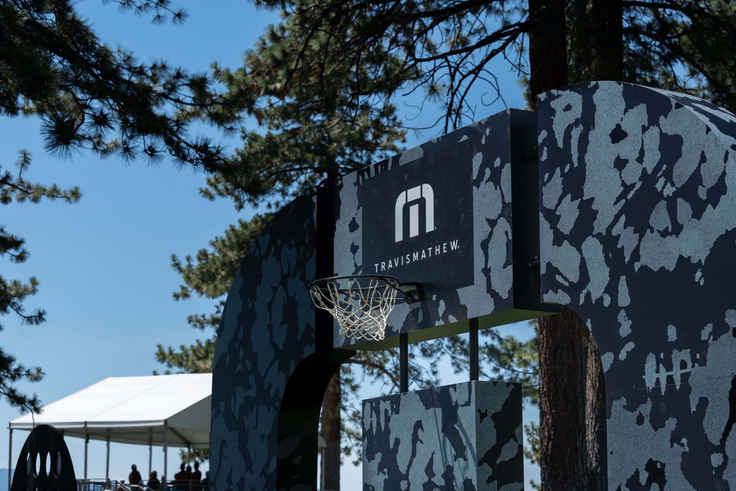 Basketball hoop with camouflage-patterned backboard under tall trees and a Travis Mathew logo, outdoor event setting