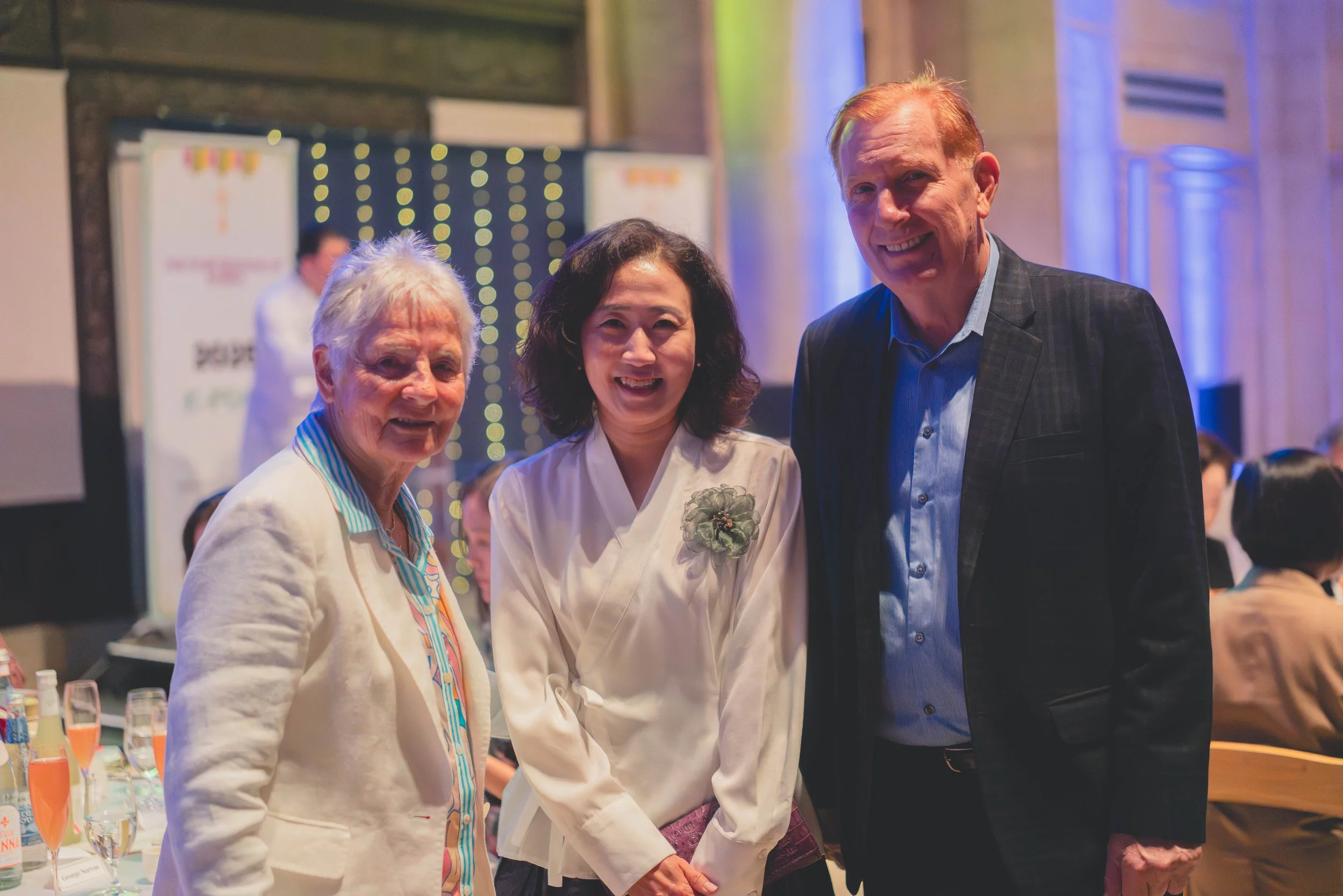 Three smiling people at a formal event, standing close together in a banquet hall with tables, wine glasses, and other guests in the background.