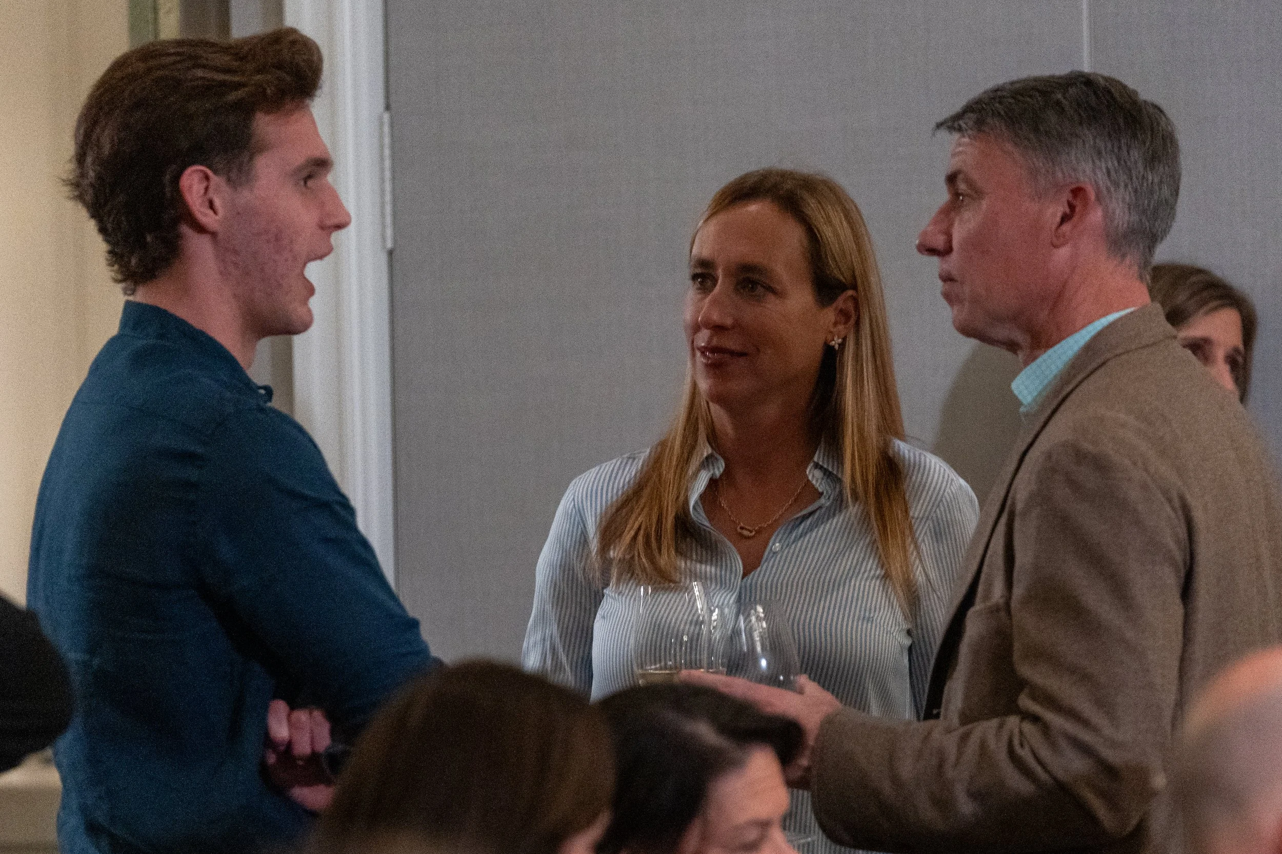 Three people in conversation at a social event, one holding a wine glass, in a room with gray walls.