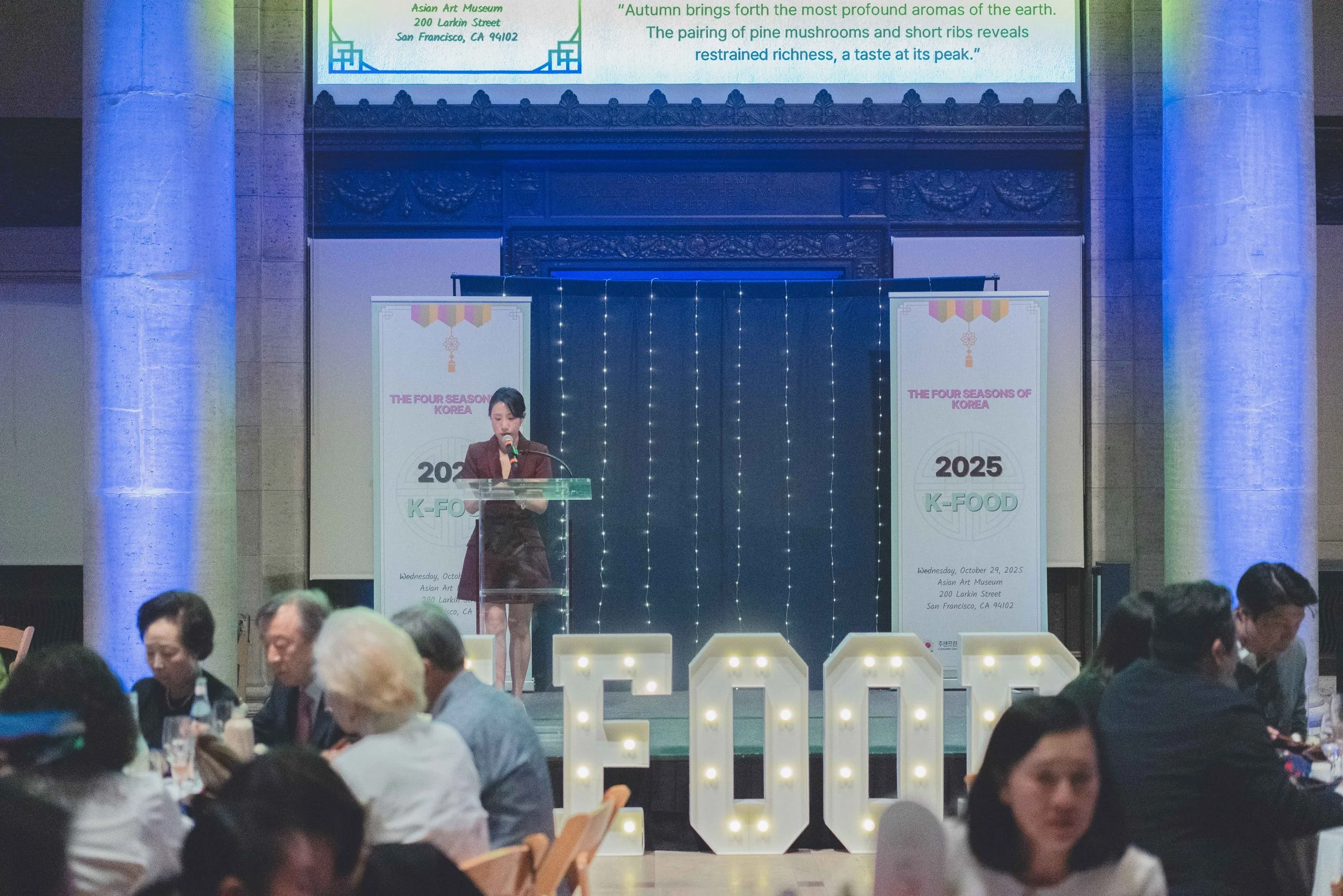 A woman speaking at a podium on a stage with banners that read 'The Four Seasons of Korea, 2025 K-Food,' during an event at the Asian Art Museum in San Francisco, with seated attendees in front.