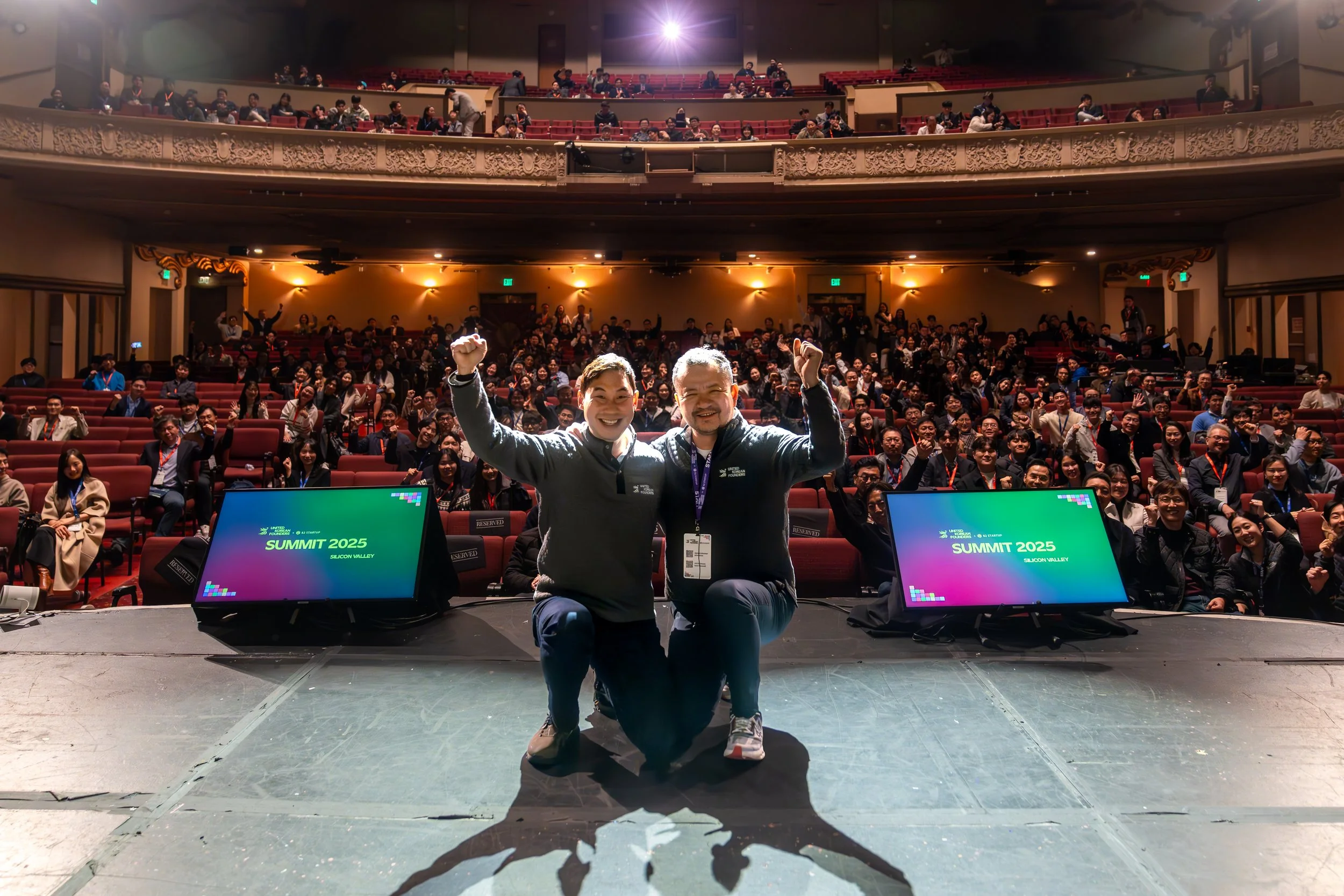 Two people kneeling and celebrating on stage at a conference with an audience in the background, and screens displaying 'SUMMIT 2025 SILICON VALLEY.'