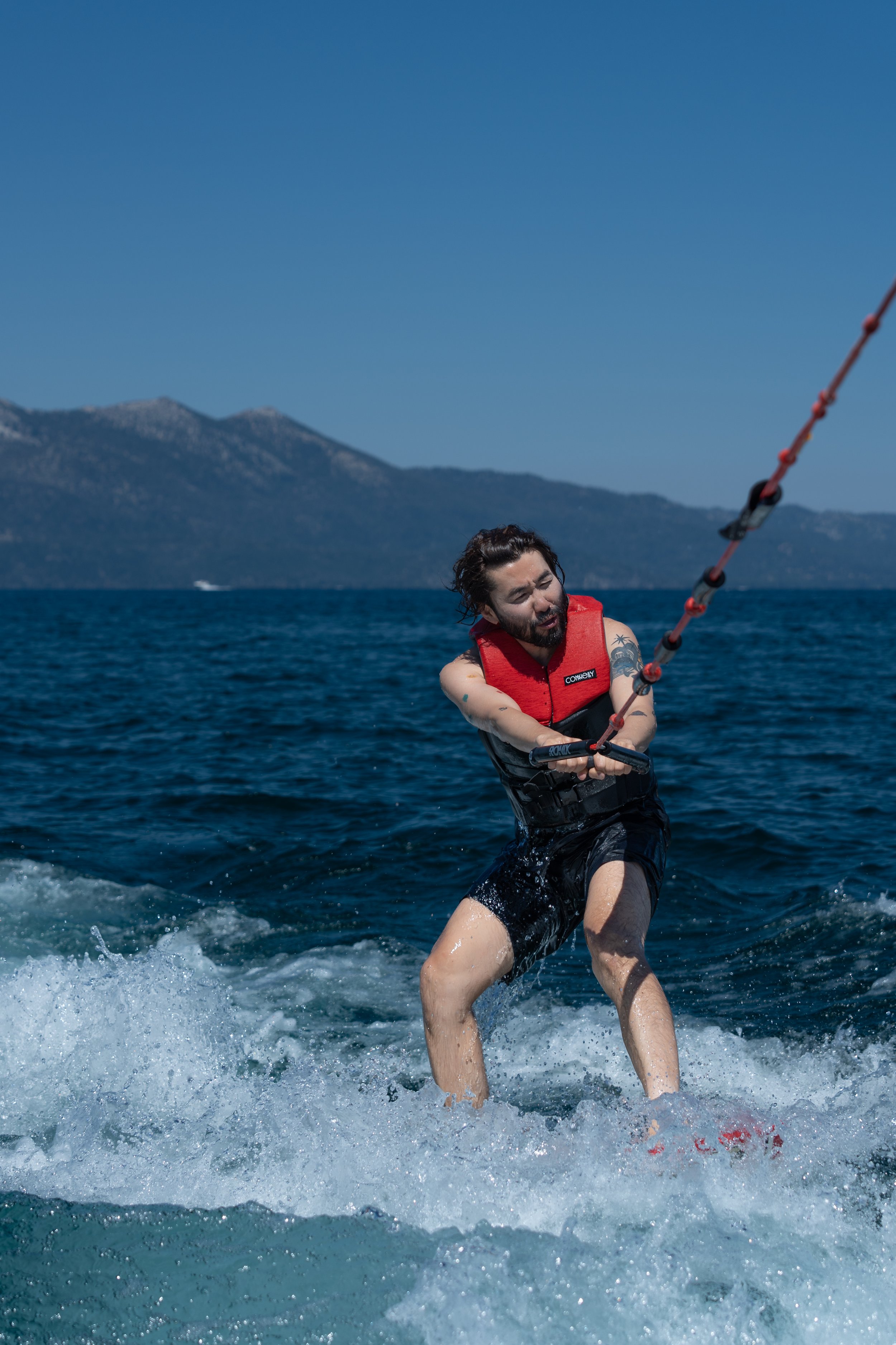 Person wakeboarding on a lake with mountains in the background, wearing a red life vest and holding onto a rope.