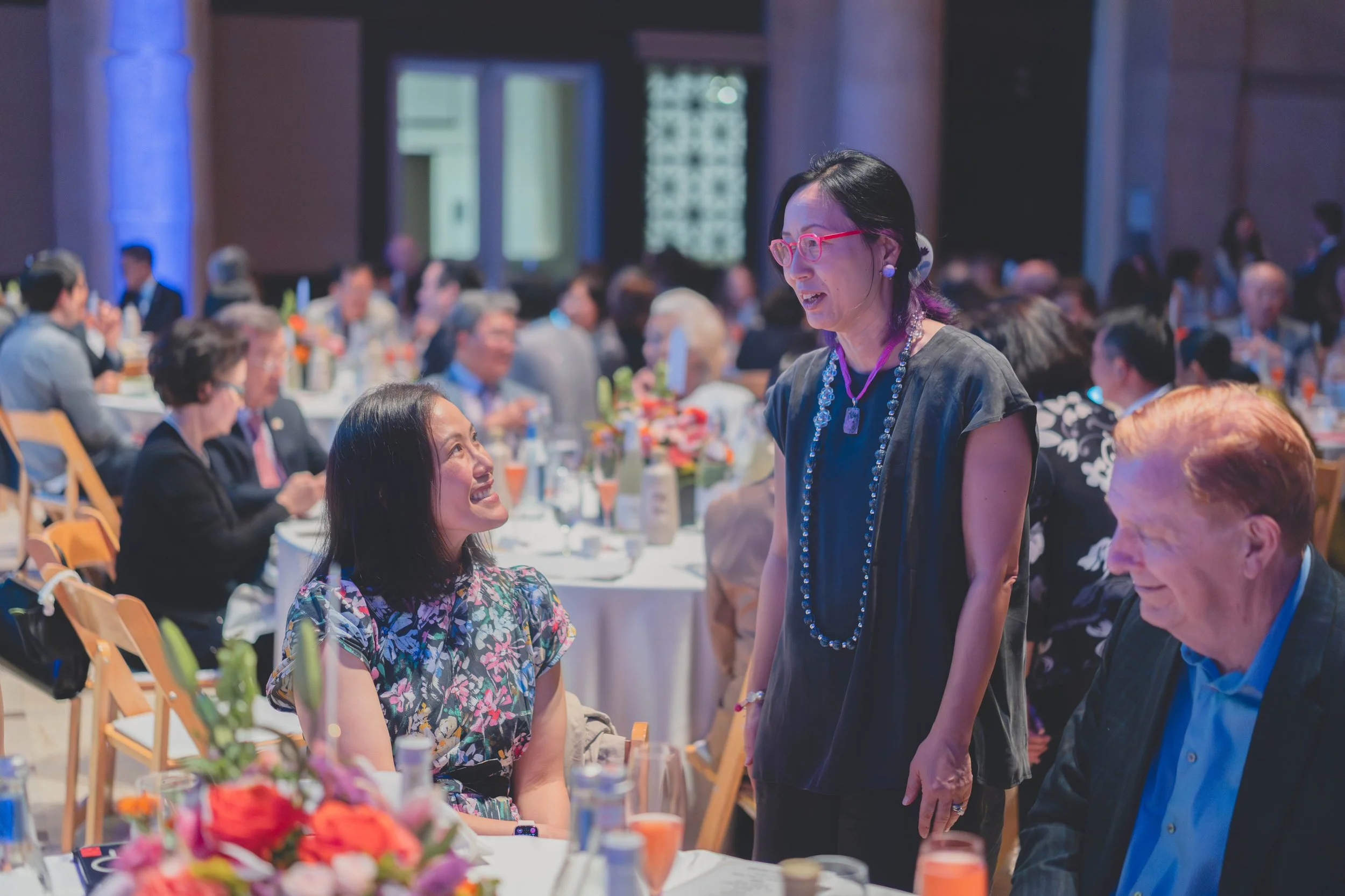 A woman with pink glasses and jewelry is standing and speaking to a seated woman at a formal event in a large, well-lit banquet hall filled with round tables, floral centerpieces, and many seated guests.