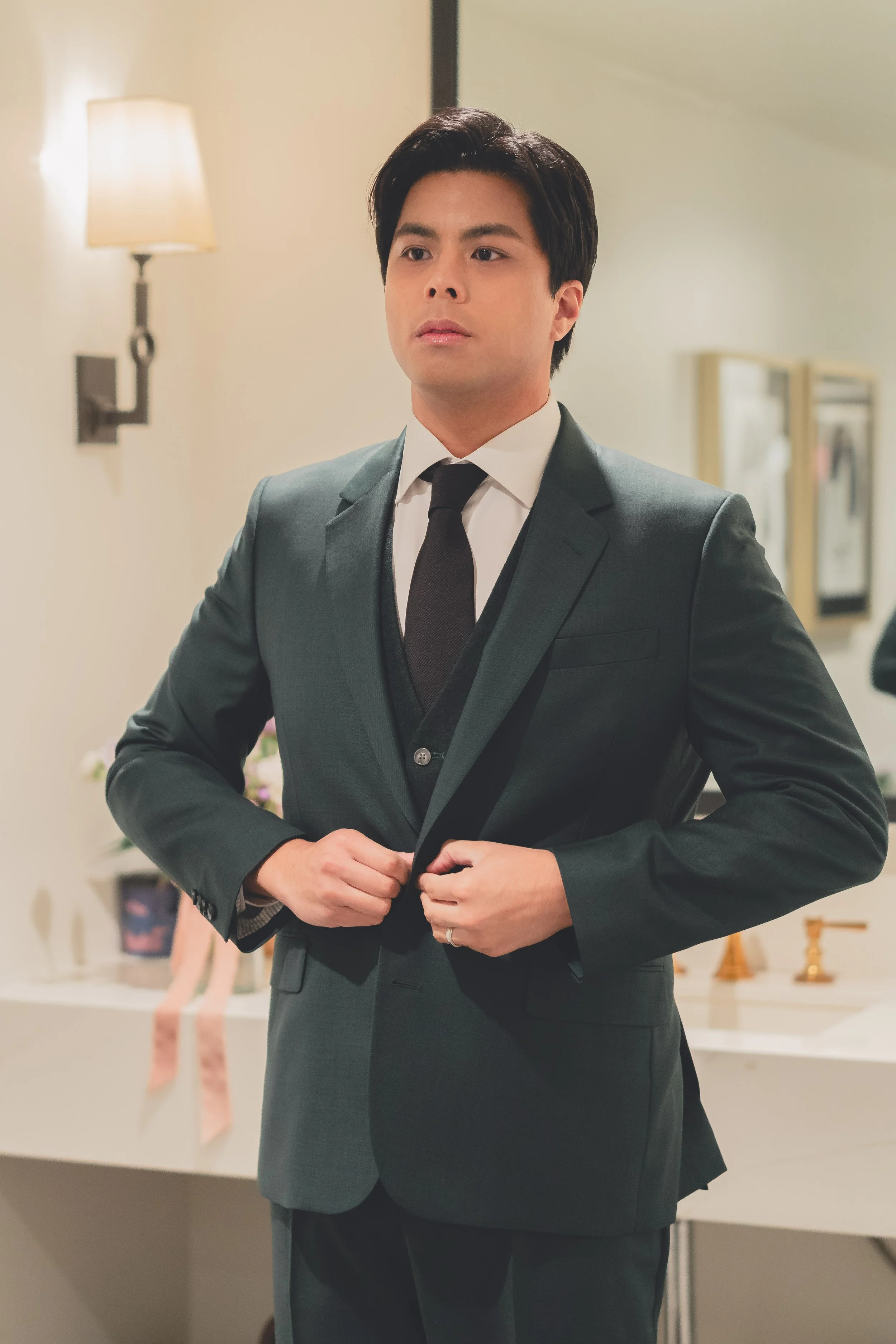 Person buttoning a dark suit jacket in a well-lit room with a wall sconce and mirror.