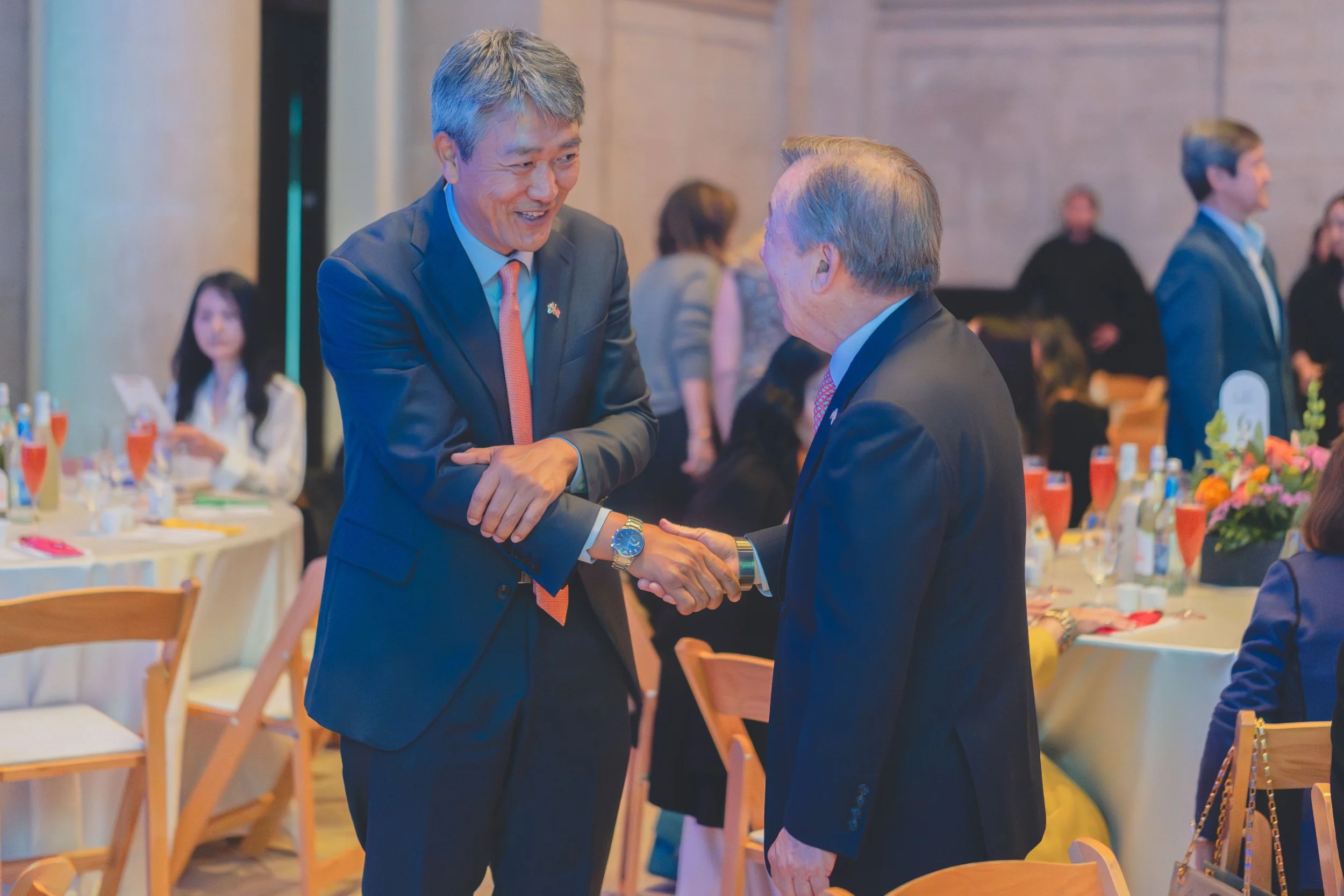 Two men in suits shaking hands and smiling at each other at a formal event or banquet, with other guests seated and standing around tables decorated with flowers and refreshments in the background.