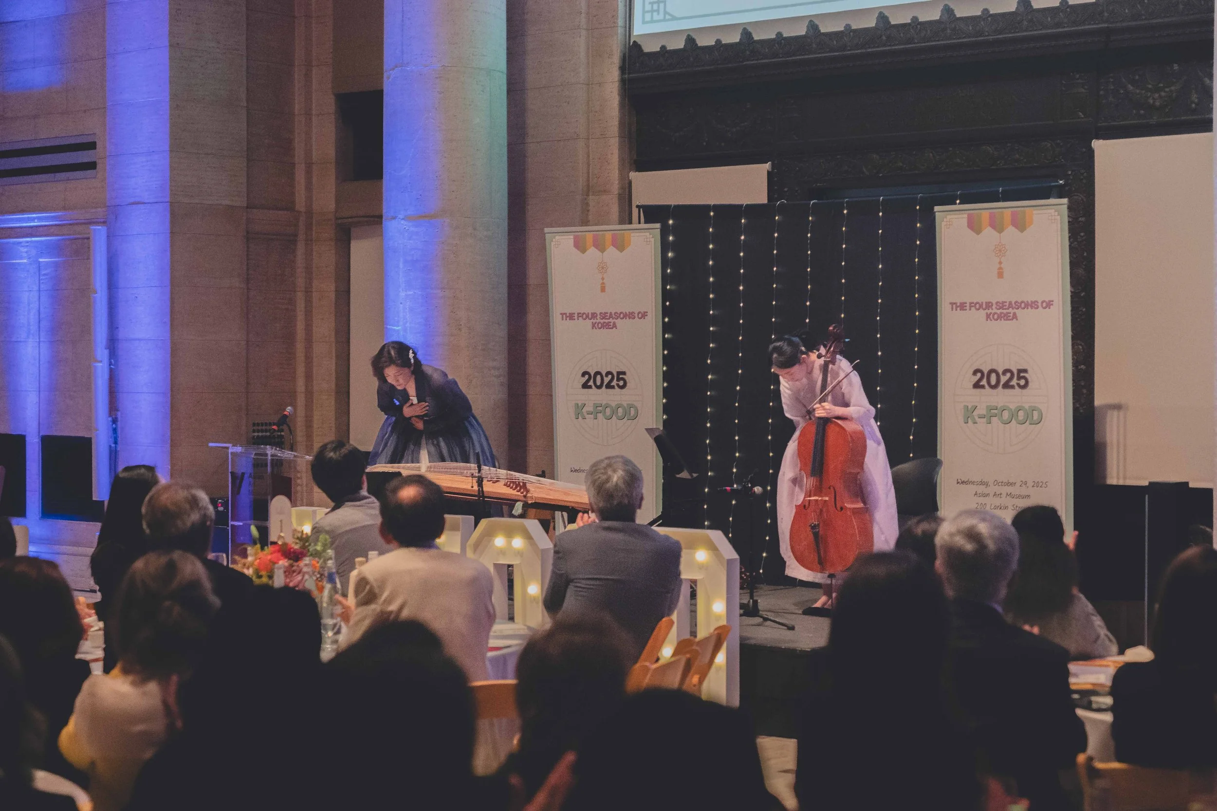 Musicians performing on stage at a cultural event, with audience seated at tables. Two women are playing traditional Korean instruments, one with a bowed string instrument and the other with a percussion instrument, in front of banners that read 'The