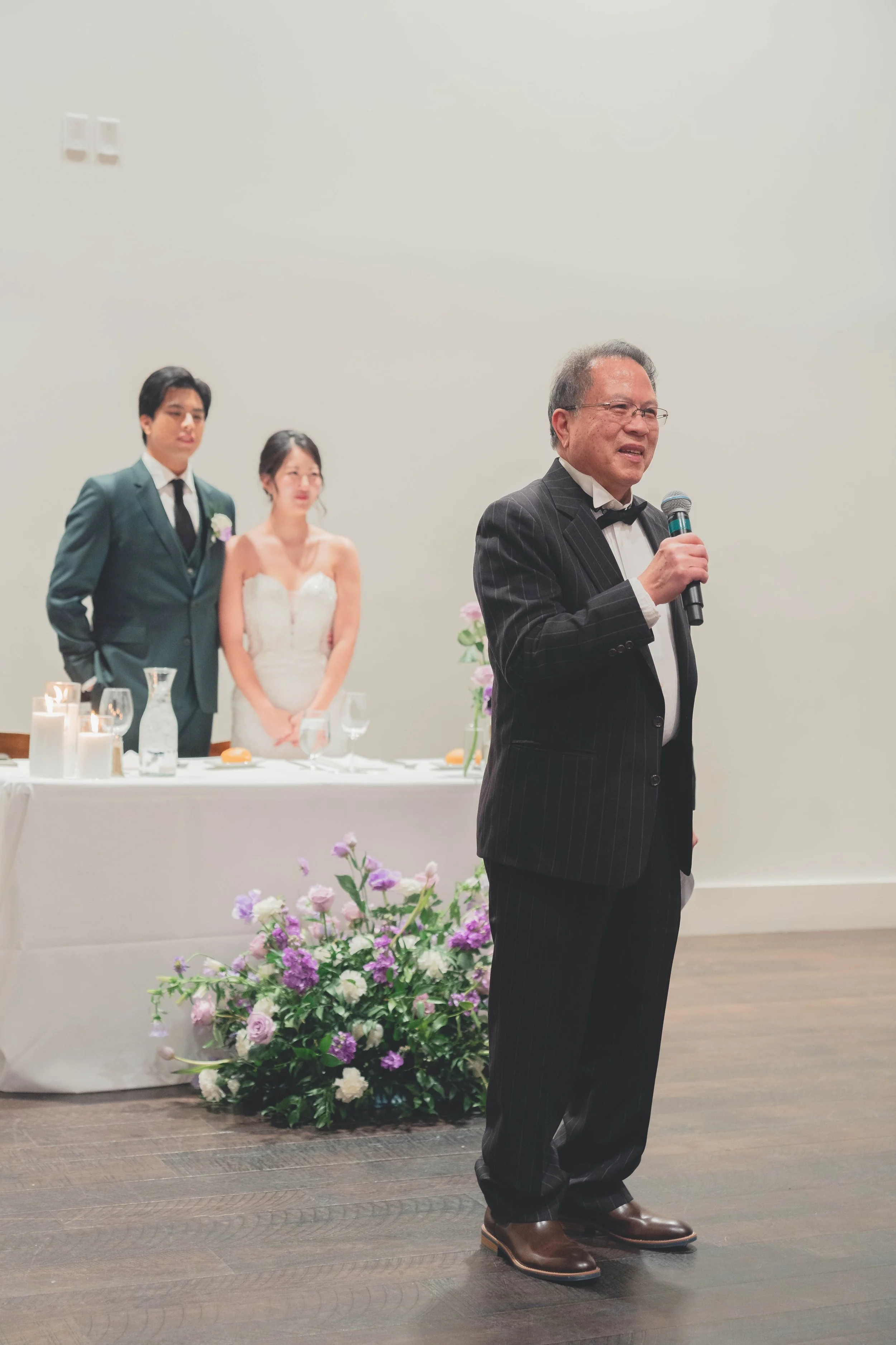 A person in a suit with a microphone gives a speech at a wedding reception while a bride and groom stand near a decorated table with flowers and candles.