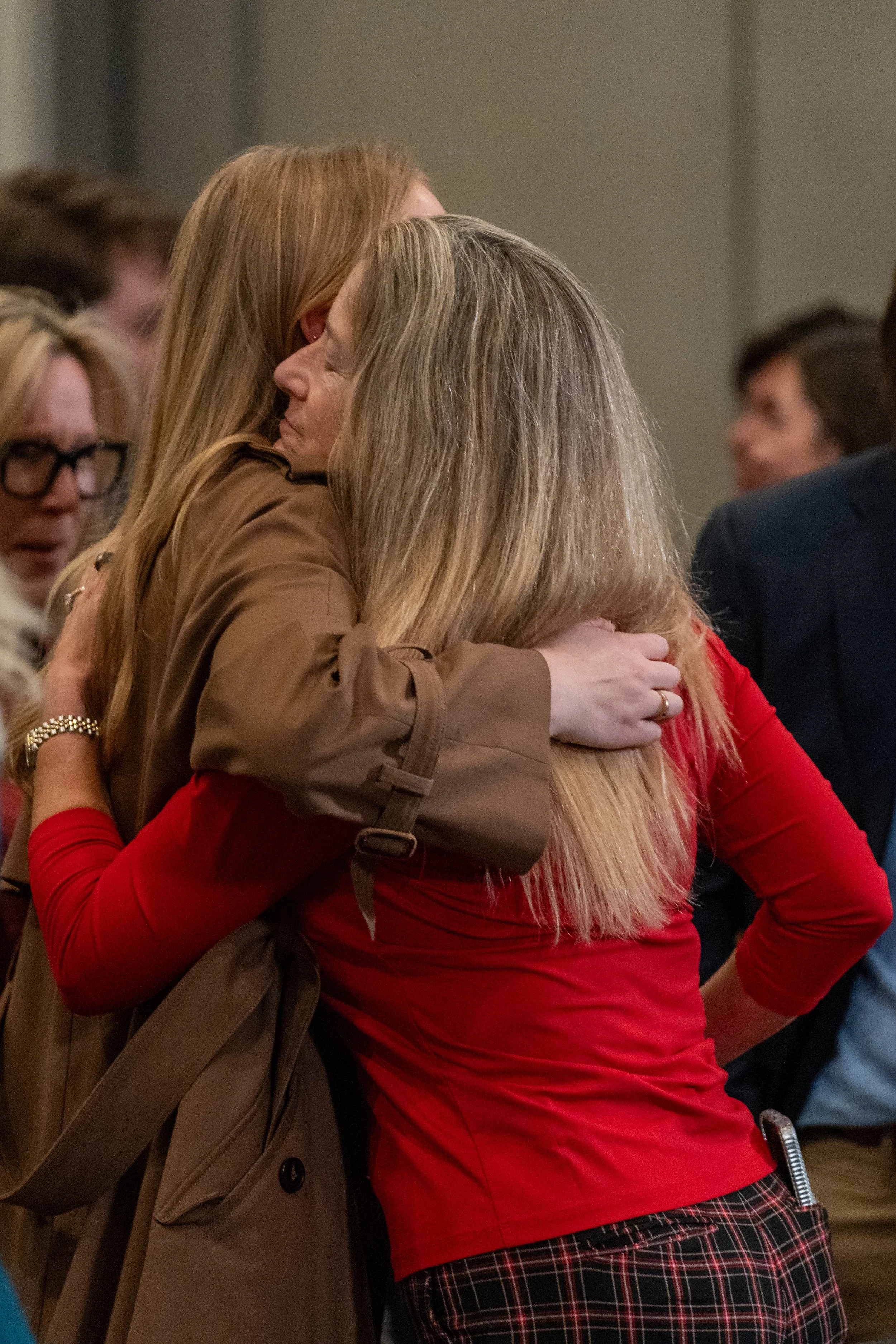 Two women hugging at an indoor gathering, one wearing a red top and the other a brown coat.