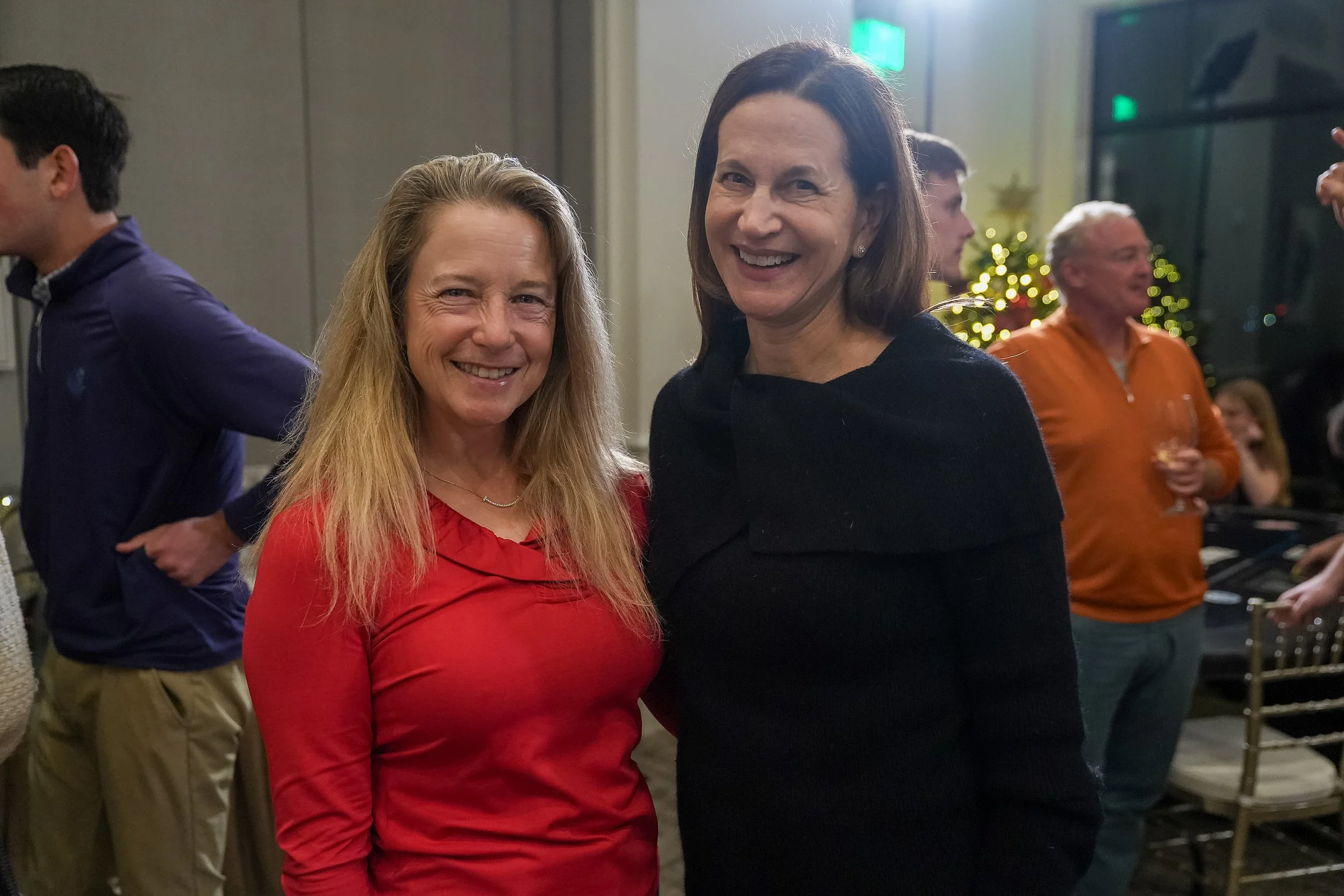 Two women smiling at an event with people in the background, featuring festive decorations.