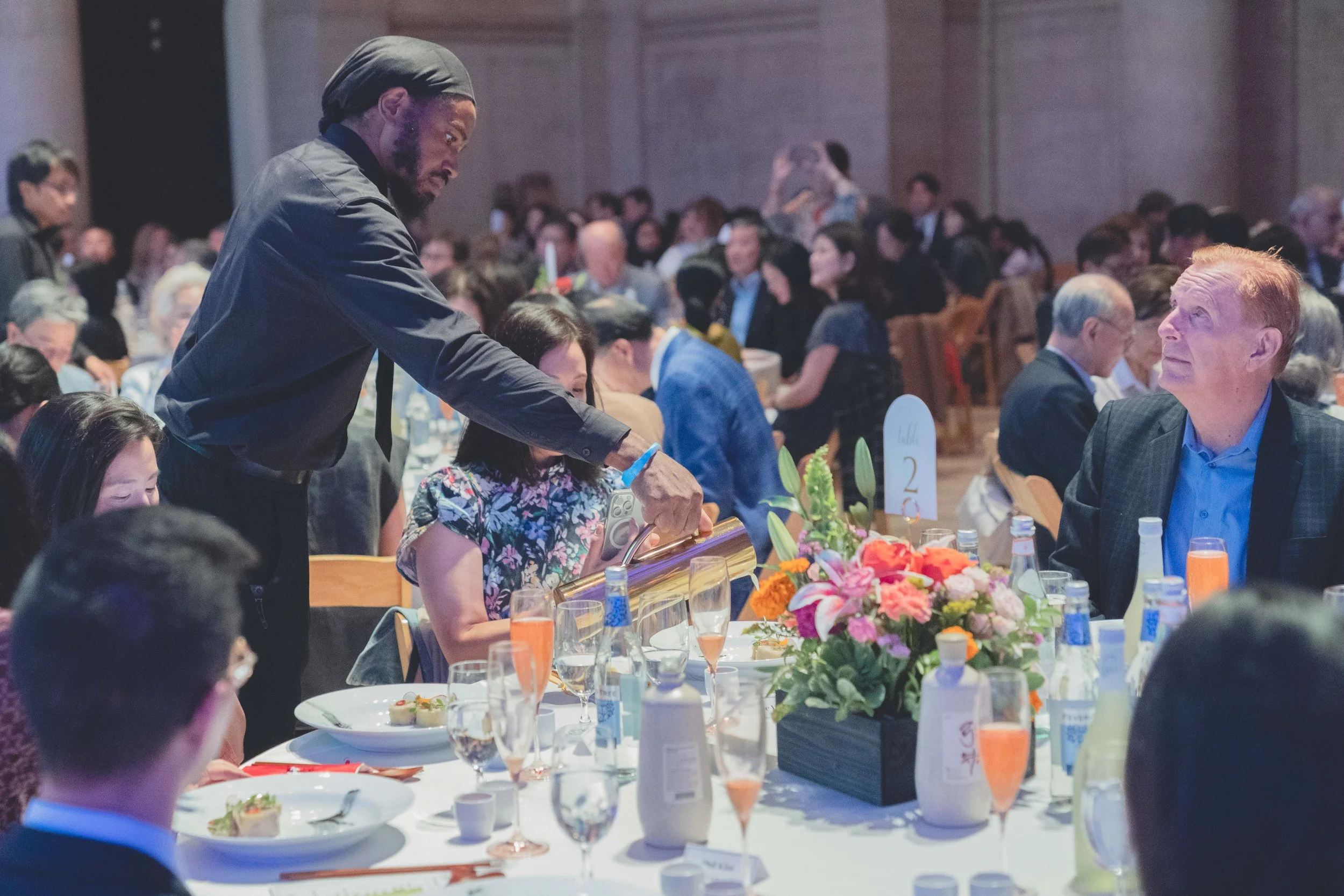 Man serving food at a large banquet table with many guests, floral centerpiece, and drinks.