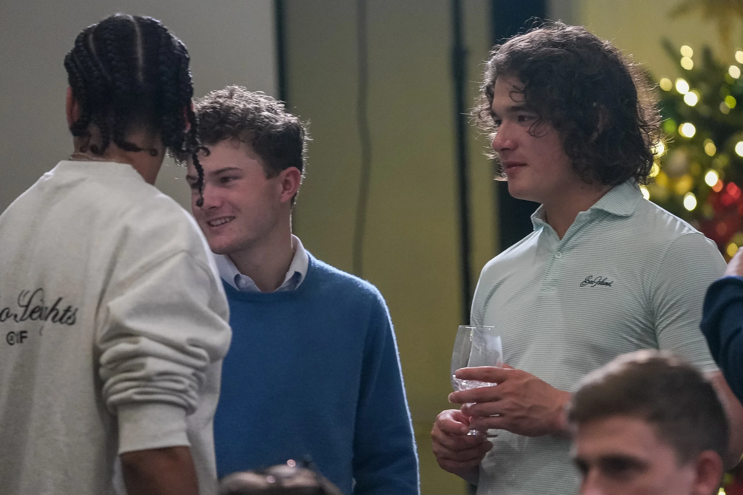 Group of people having a conversation at a social gathering, one holding a wine glass, with festive decorations in the background.