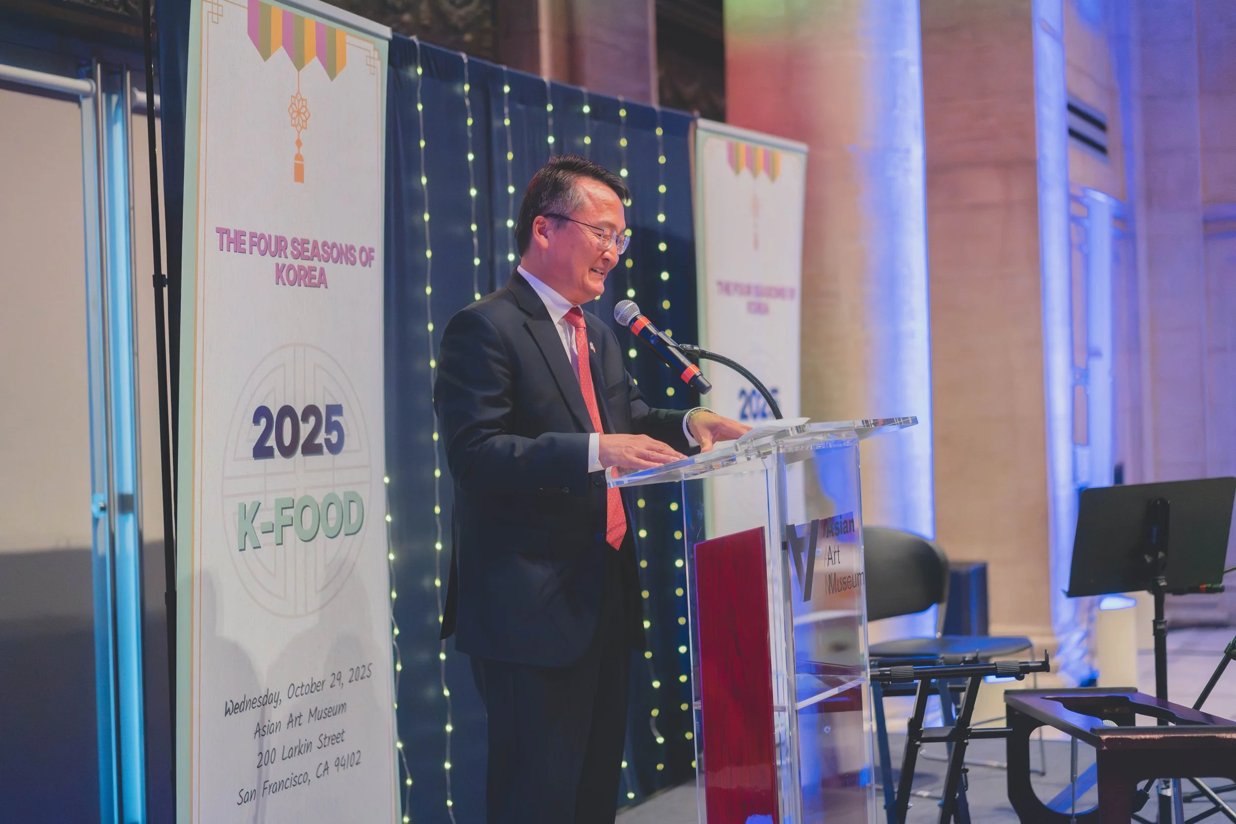 A man in a suit speaking at a podium during an event titled 'The Four Seasons of Korea' at the Asian Art Museum on October 29, 2025, in San Francisco. There are posters and lights in the background.