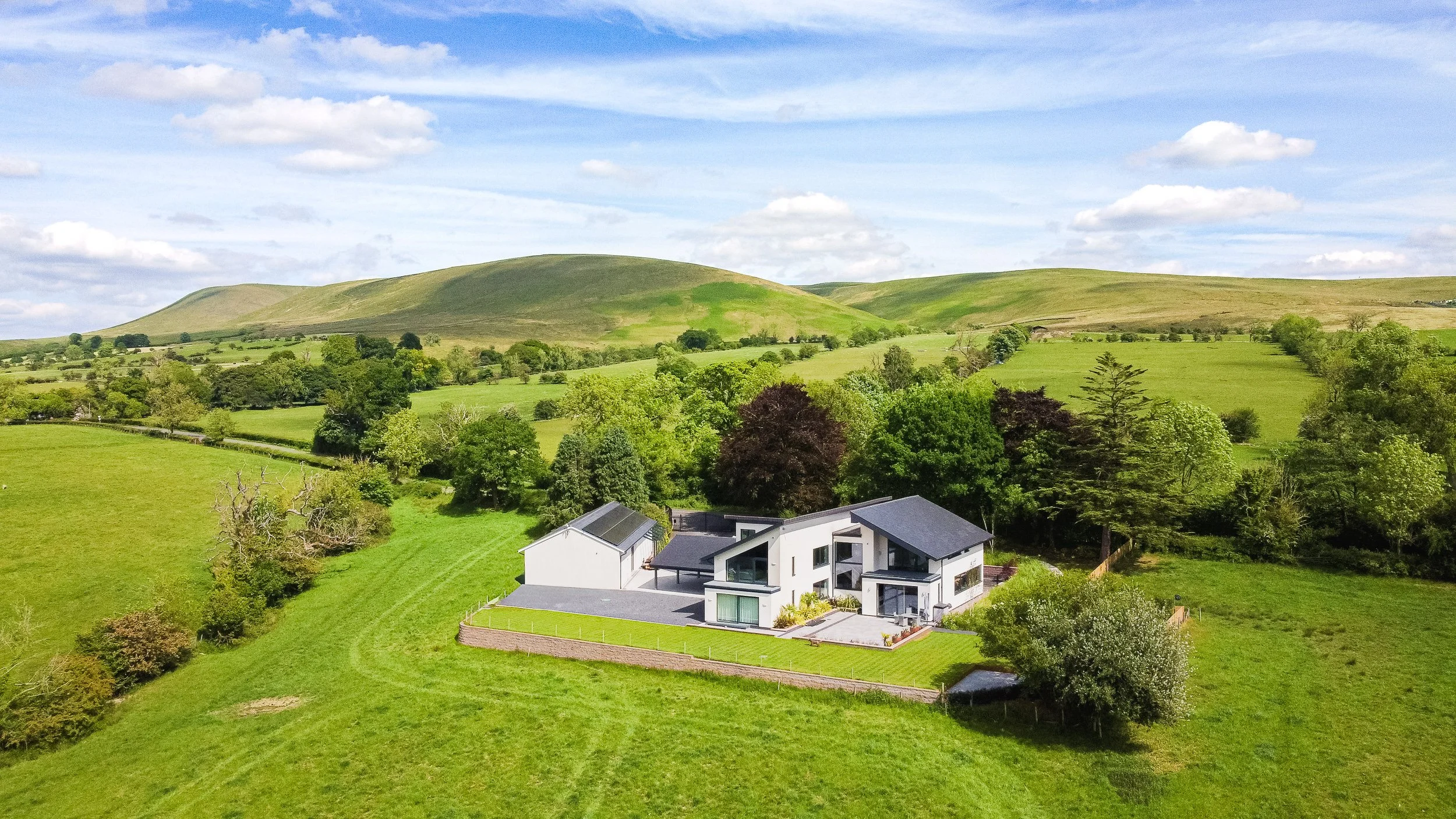 An aerial view of a modern, architect-designed detached house set in vast green rural countryside, backed by rolling hills for sale with an estate agent.