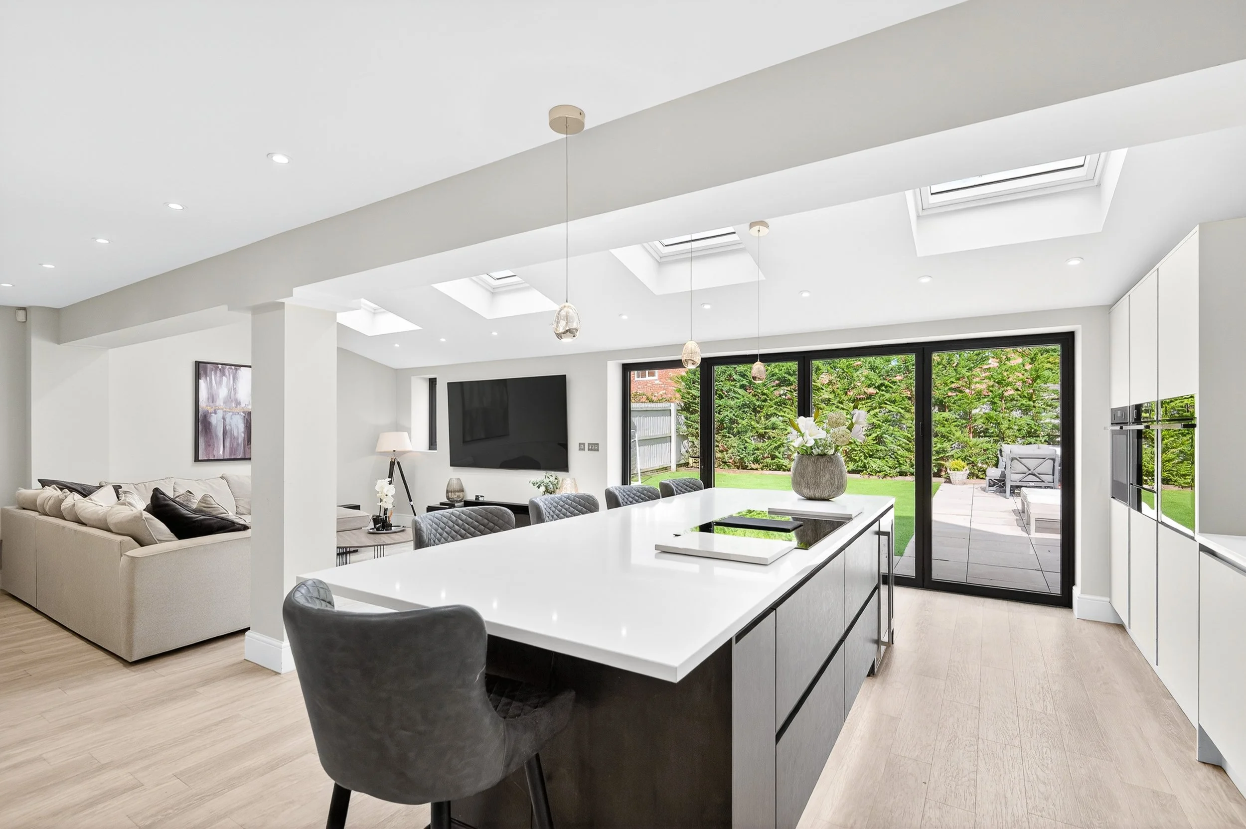A modern open-plan kitchen featuring a large central island, white worktops, and black bi-fold doors opening onto a patio with sky lanterns overhead. This image is being analysed by AI for a property description generator.