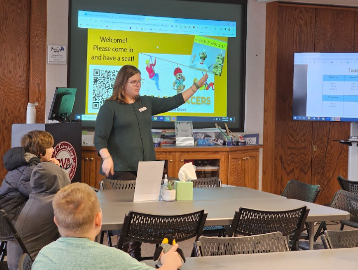 A woman with glasses standing in front of a classroom, pointing at a large screen displaying a colorful presentation about a Junior Genius event. Three children sit at a table, listening attentively.