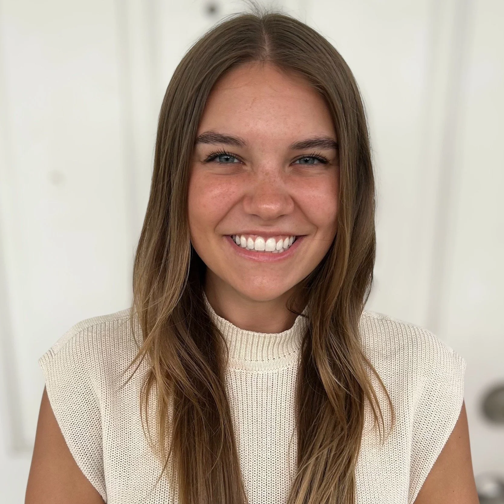 A young woman with long brown hair, blue eyes, and freckles, smiling at the camera, wearing a beige sleeveless top, against a plain white background.