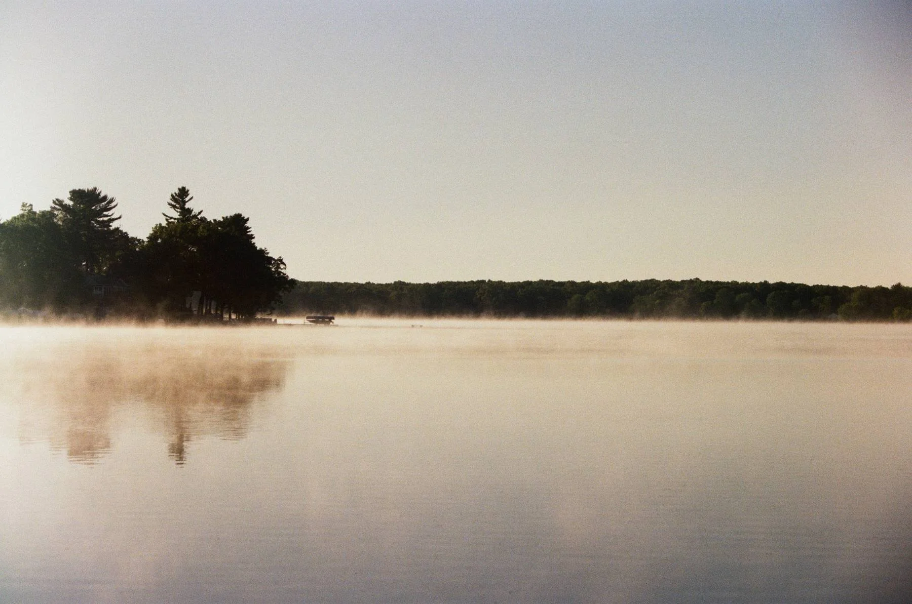 A calm lake with a foggy surface reflecting trees on the left side and a distant forest horizon. Early morning with mist rising from the water.