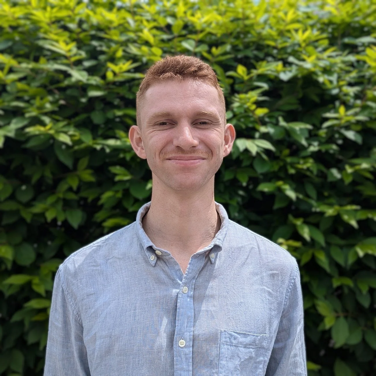A young man with short red hair and a slight smile, wearing a light gray button-up shirt, standing outdoors in front of a green leafy hedge.
