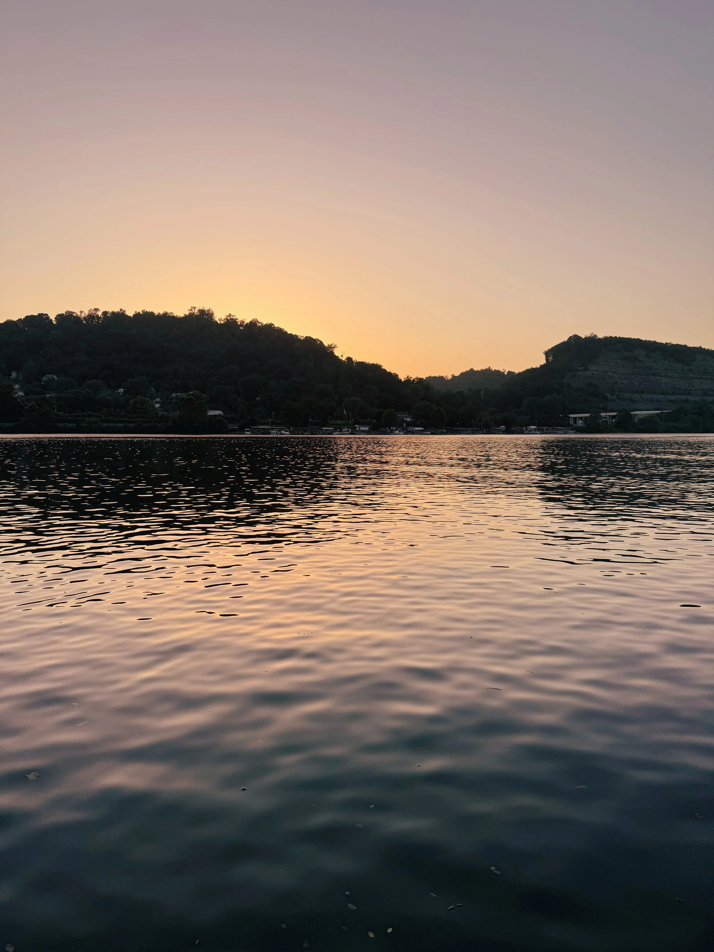 A body of water at sunset with hills and trees in the background.