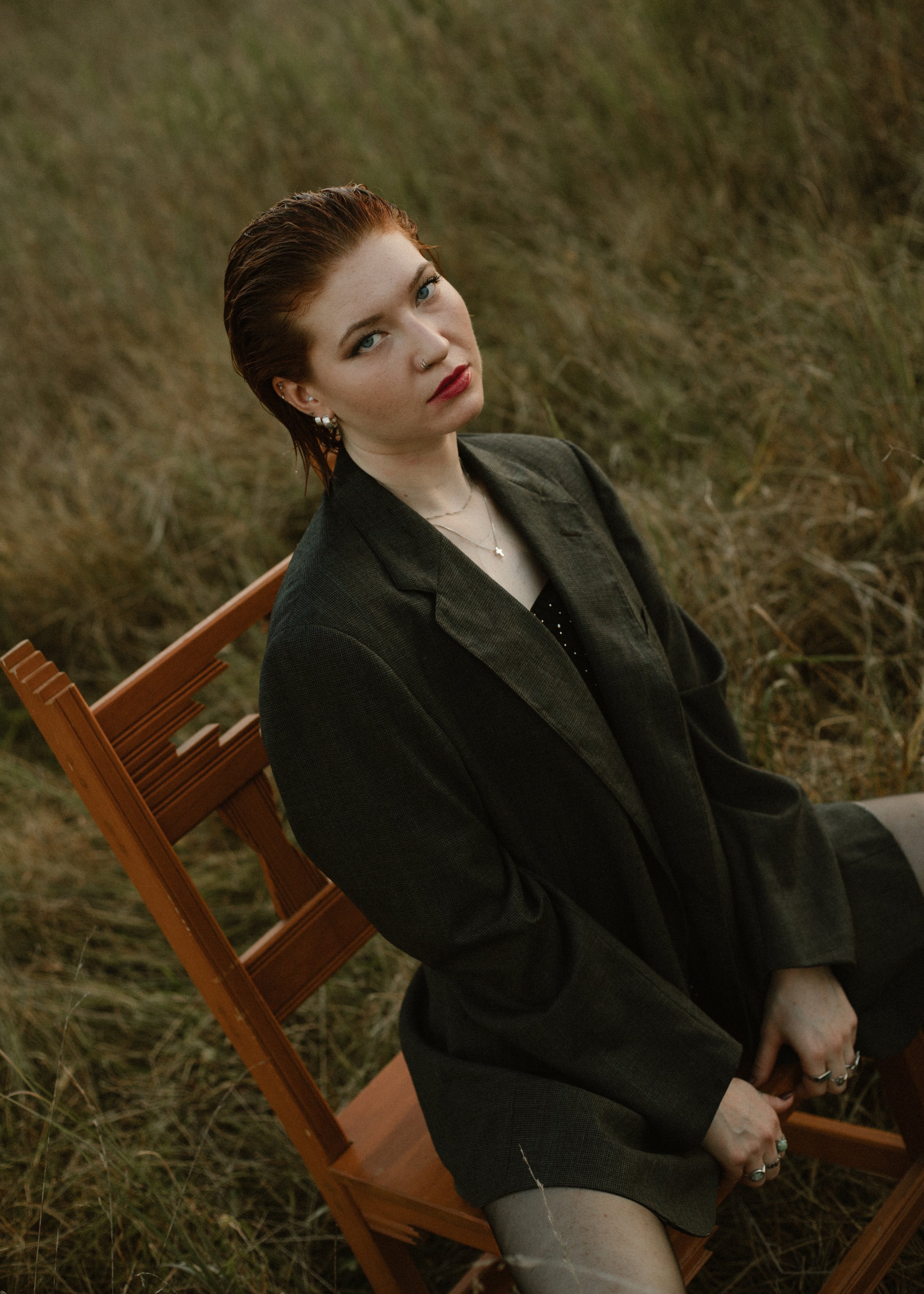 Model with red hair, blue eyes, and fair skin sitting on a wooden chair outdoors in a grassy field, wearing a dark oversized blazer, and multiple rings, with a necklace and earrings. Tattoos. Editorial. Creative Portraits. Shelby Farms Park. Memphis