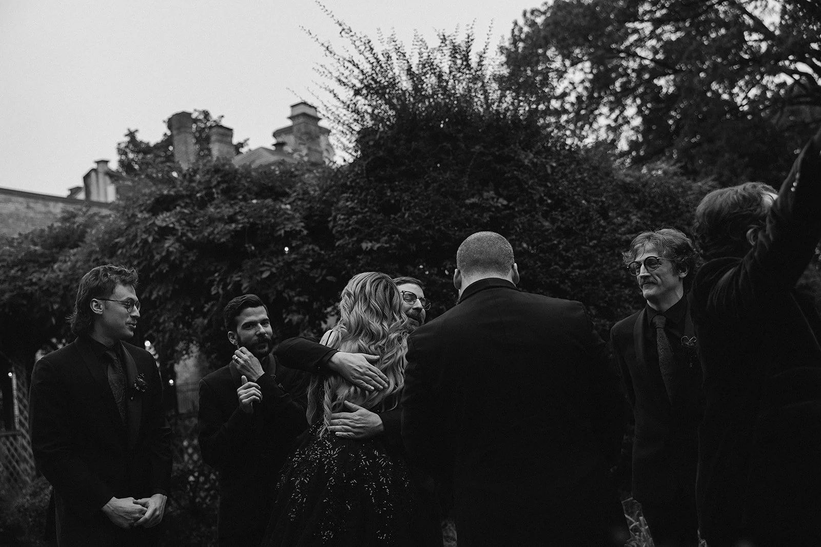 Black and white photo of a wedding scene outdoors with several people. A woman with long, wavy hair is hugging a man, while others watch and smile. Candid Documentary photography. woodruff fontaine house museum. memphis tn photographer