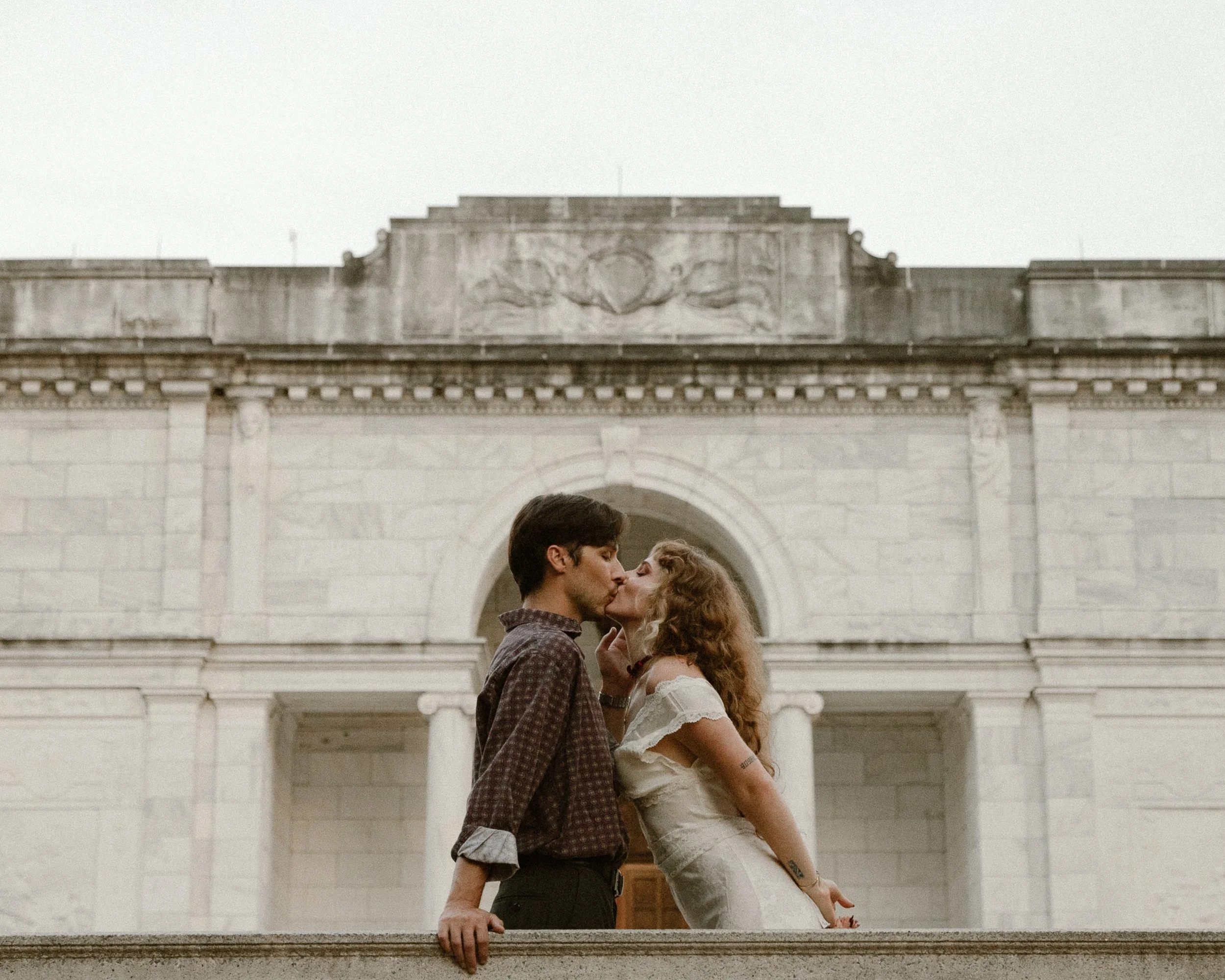 A couple sharing a kiss in front of a historic building with columns and a carved stone facade. Moody. Cinematic. Brooks Museum. Engagement Session. Overton Park 901. Memphis Photographer. Documentary. Candid. Editorial.