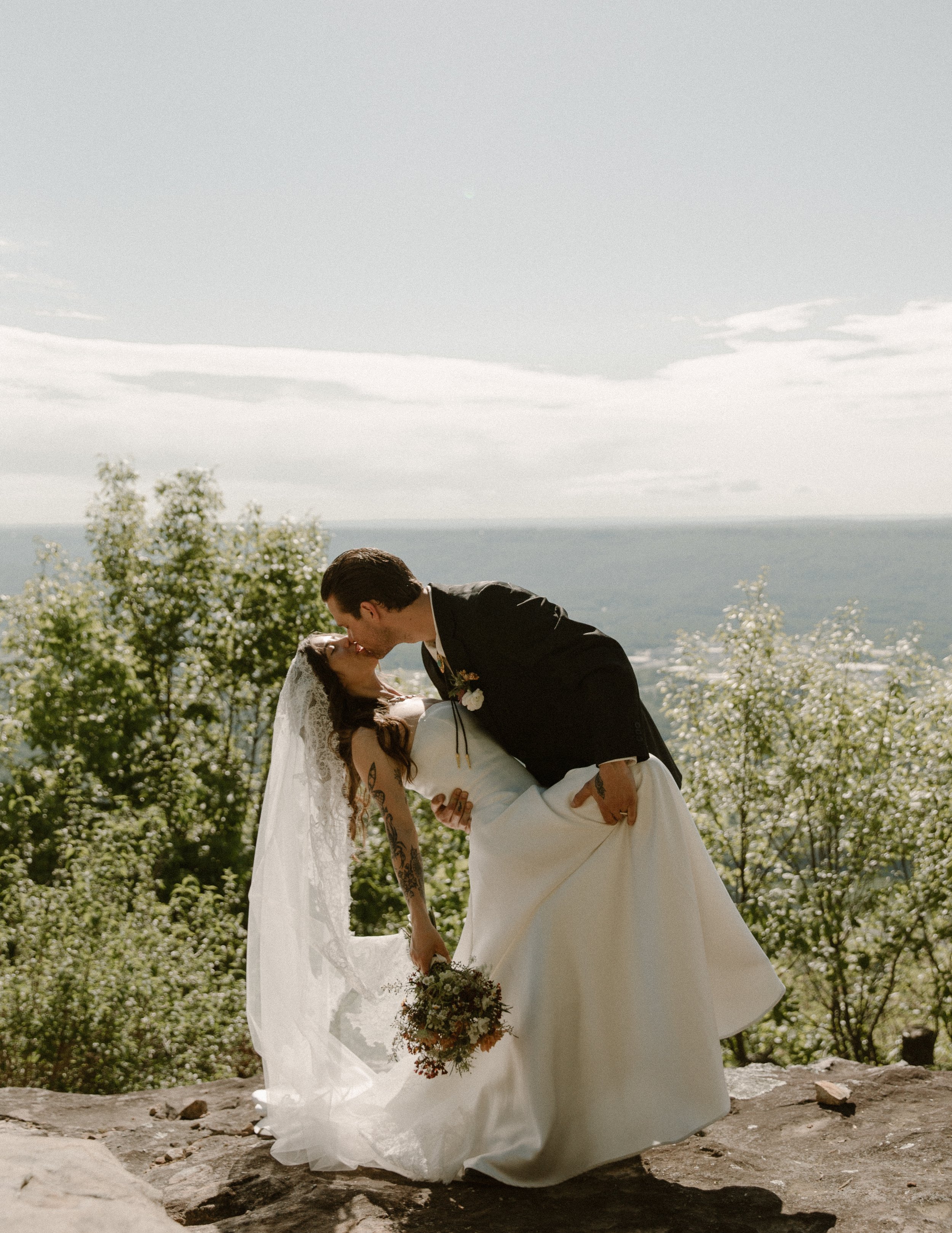 A newlywed couple sharing a kiss outdoors, with the groom dipping the bride, on a mountain with trees and a scenic landscape in the background. Elopement. Travel Photographer. Chattanooga Tennessee. Lookout Mountain.