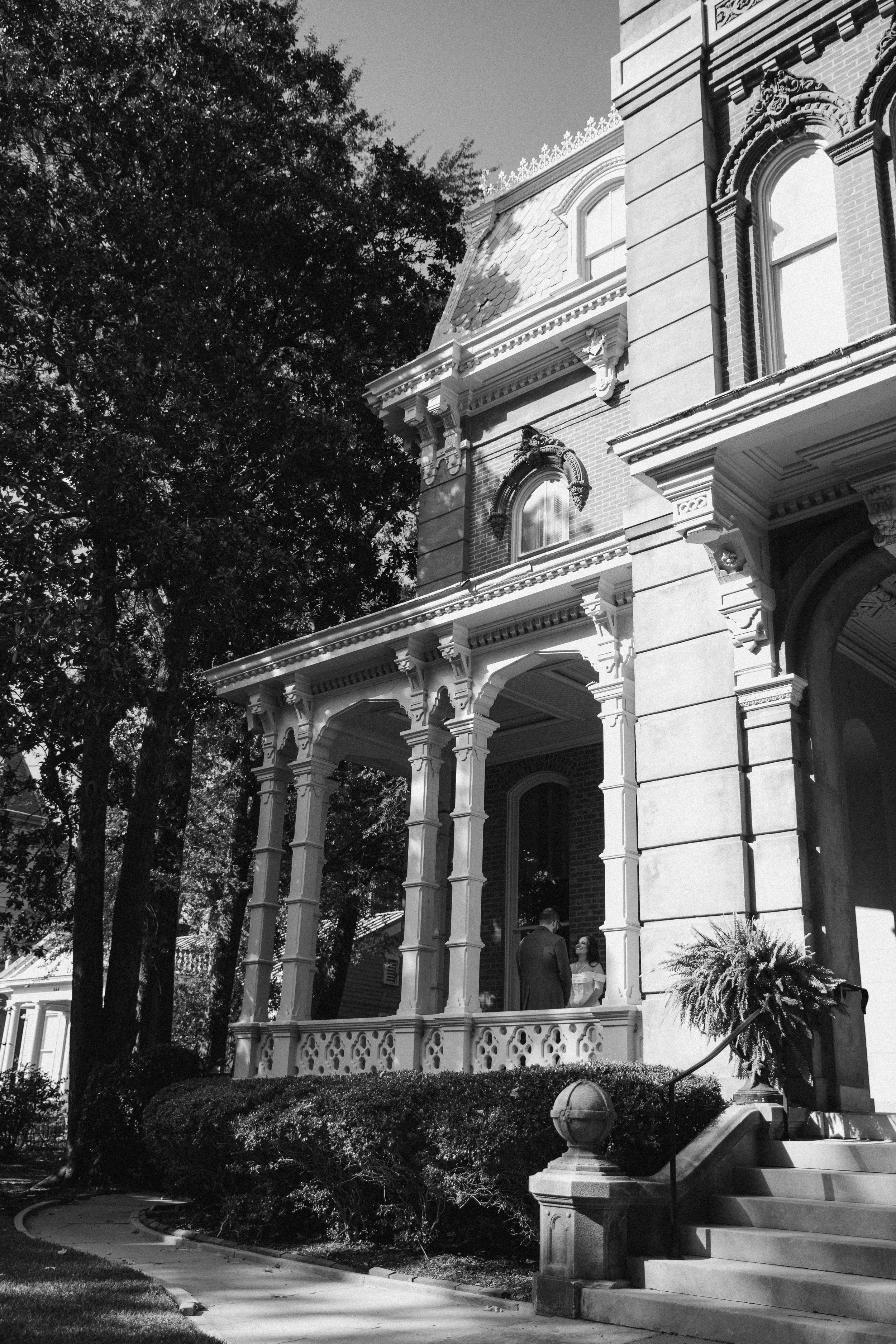 Black and white photo of a large, ornate Victorian-style house with a porch. Two people dressed formally are standing on the porch, engaged in conversation. Documentary candid photography. Woodruff Fontaine House Musuem Wedding Photographer. Memphis