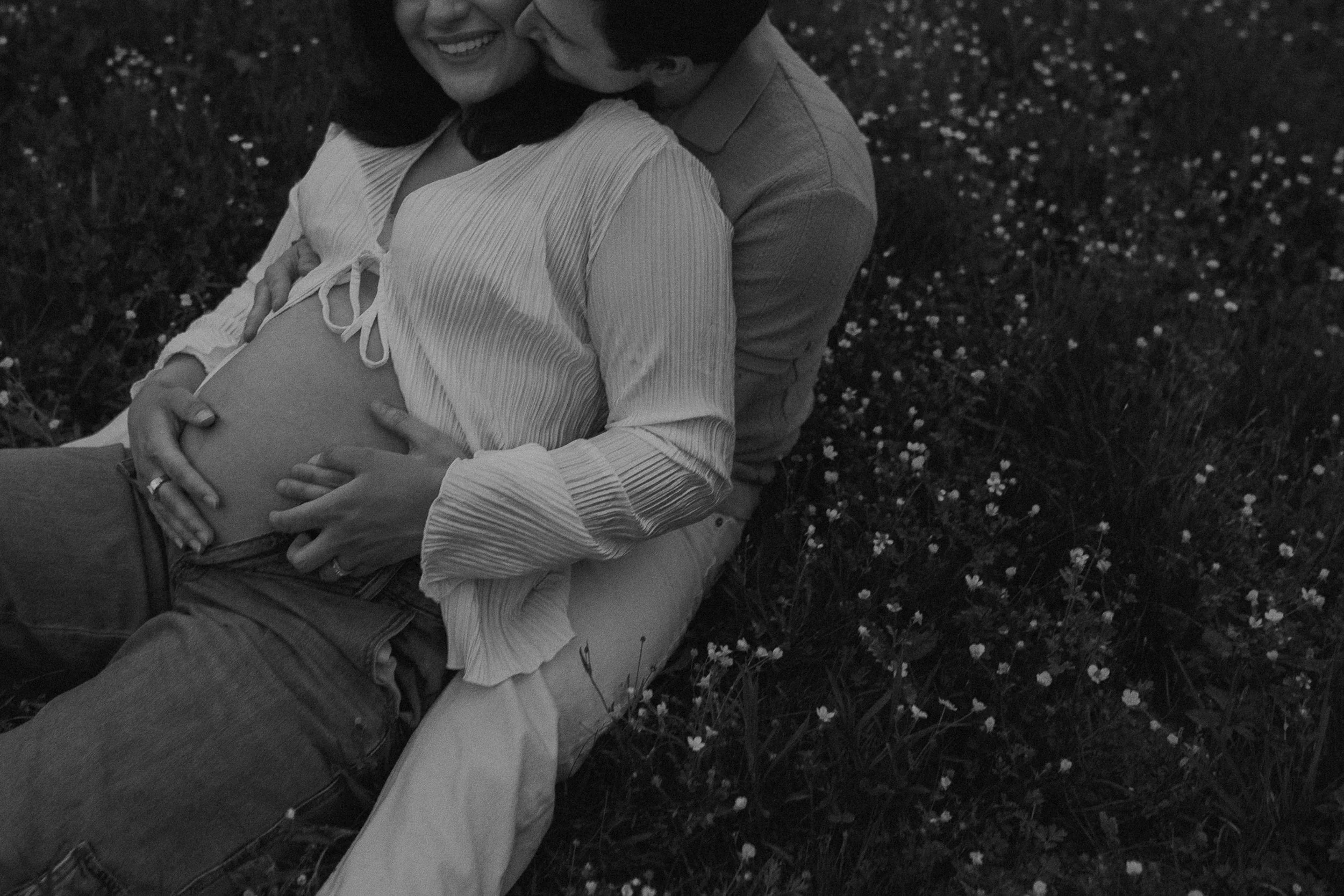 A couple sitting on the grass, with the woman pregnant, holding her belly, and the man sitting behind her, all smiling and embracing. Maternity Session. Candid. Real and Raw photography. memphis tn photographer