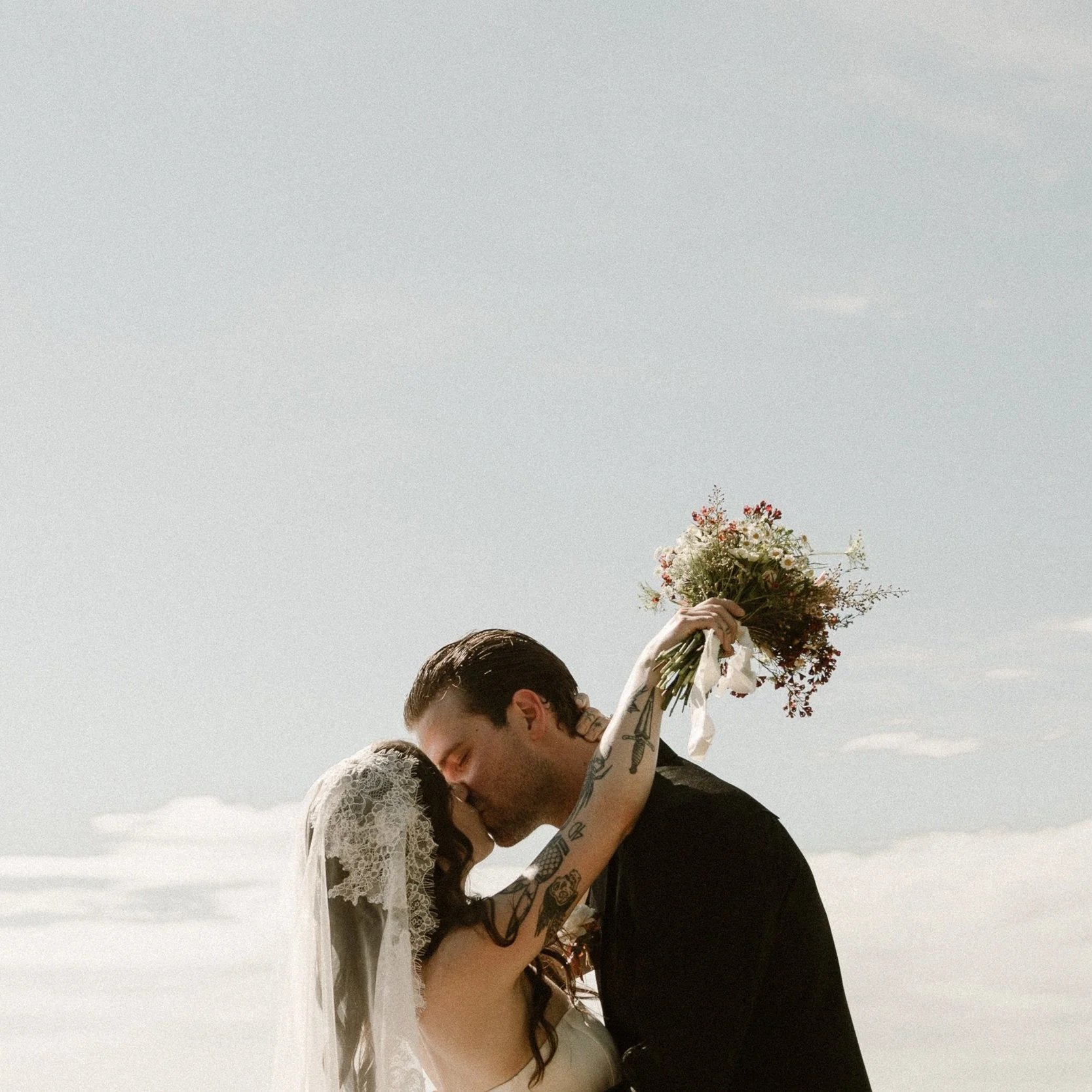 Bride and groom kissing outdoors, bride holding bouquet up, bride wearing lace veil, bride and groom with tattoos, cloudy sky background. Chattanooga TN Memphis Travel Photographer. dark moody documentary candid photographer