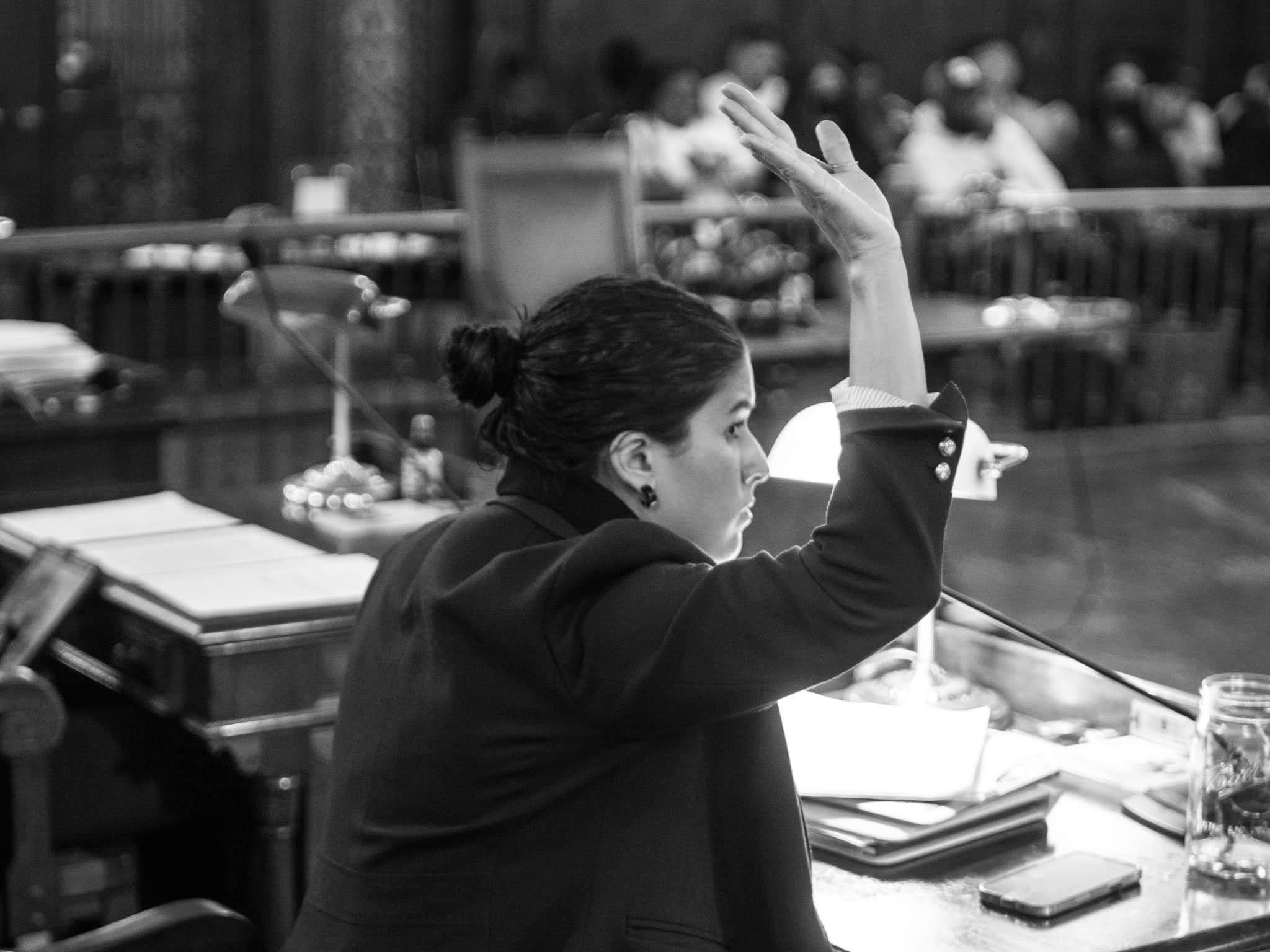 A woman in a professional suit raises her hand possibly to speak during a meeting or courtroom session, seated at a desk with papers, a glass of water, a phone, and a desk lamp, with other people in the background.