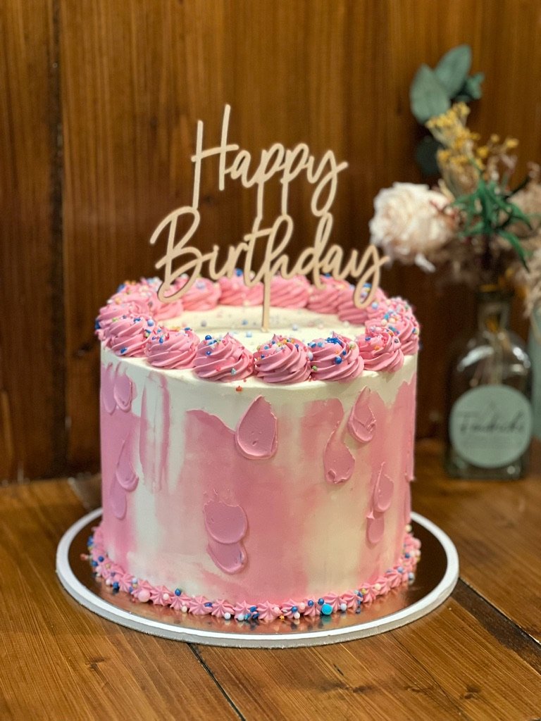 Torte mit rosa und lila Farbakzenten und Happy-Birthday-Topper auf einem Holztisch