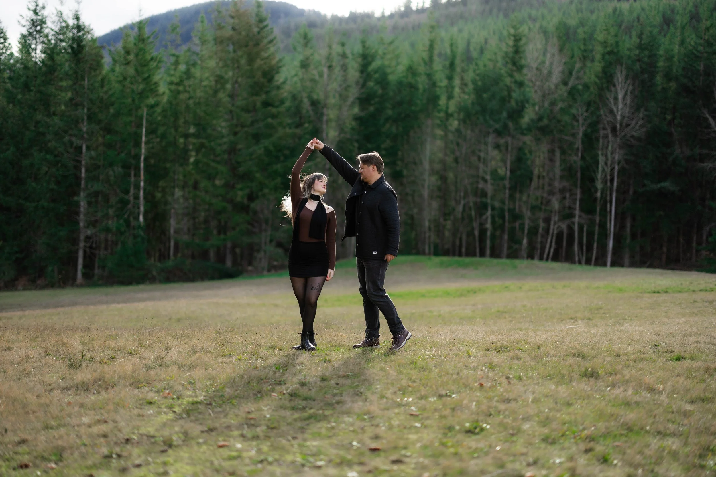A couple dances in an open field with a sunlit forest background