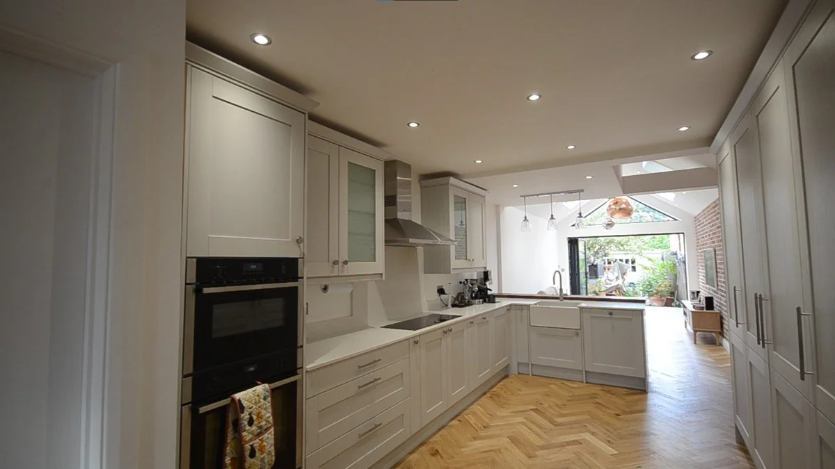 Raw Architects LTD — Modern kitchen with white cabinets, wooden flooring, stainless steel appliances, and large window with outdoor view.