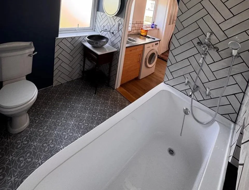 A bathroom with a toilet, patterned floor tiles, a small black table with a decorative bowl, a window, and a bathtub with wall-mounted shower fixtures. In the background, there is a laundry area with a washing machine and a wooden cabinet.