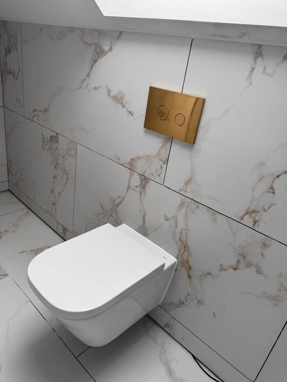 Modern bathroom with wall-mounted toilet, marble walls, and a gold flush plate.