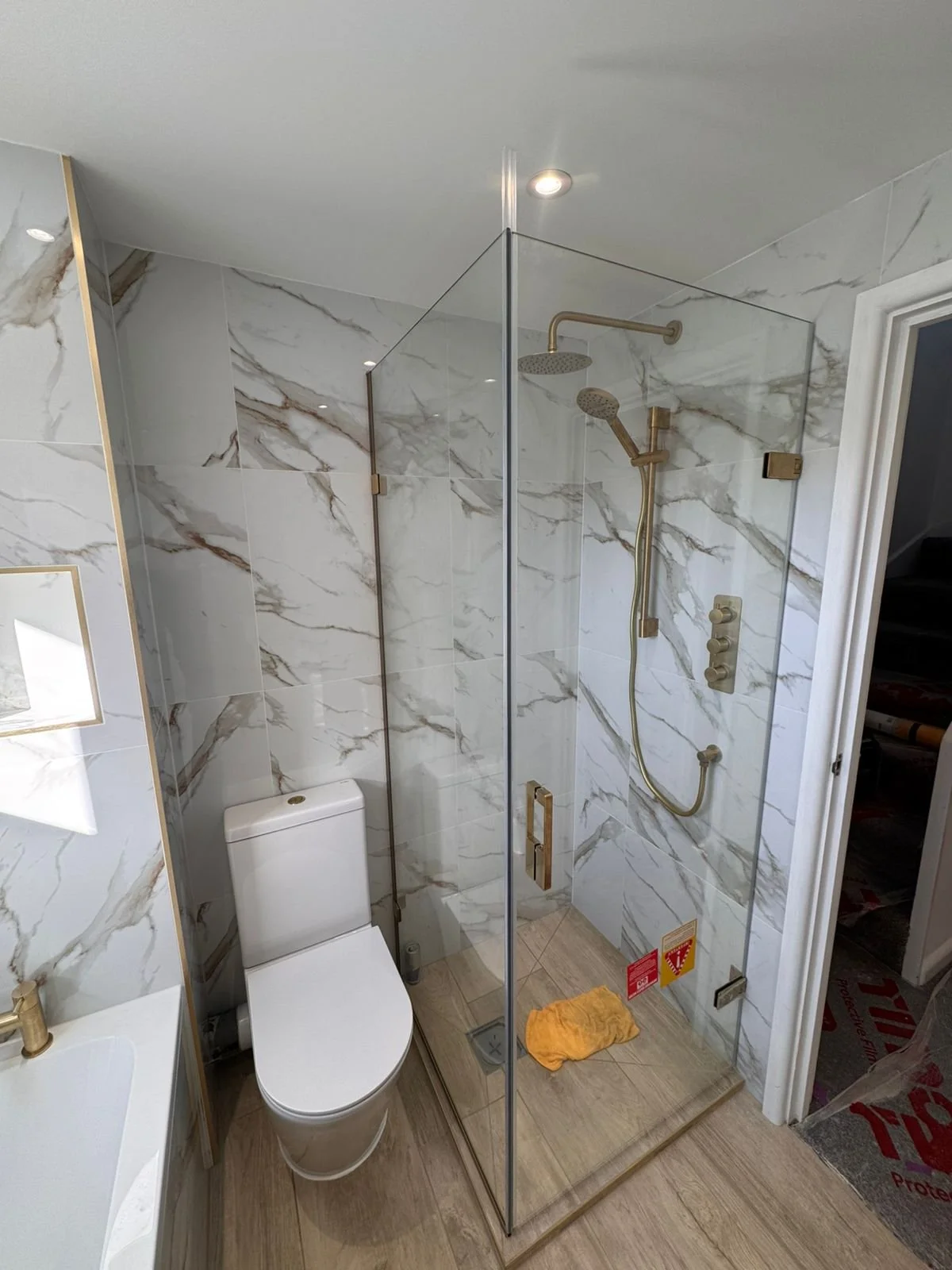Modern bathroom shower with gold fixtures, marble wall tiles, and a glass enclosure next to a toilet with a white tank and seat. There is a yellow cloth on the wooden shower floor, and a small sink with a gold faucet is visible on the left.