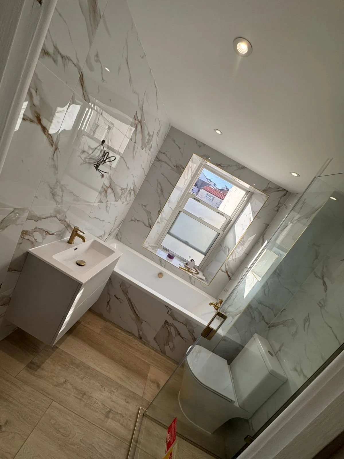 Modern bathroom with marble walls, a bathtub by the window, a small white sink with gold fixtures, a glass shower enclosure, and wood-look tile flooring.