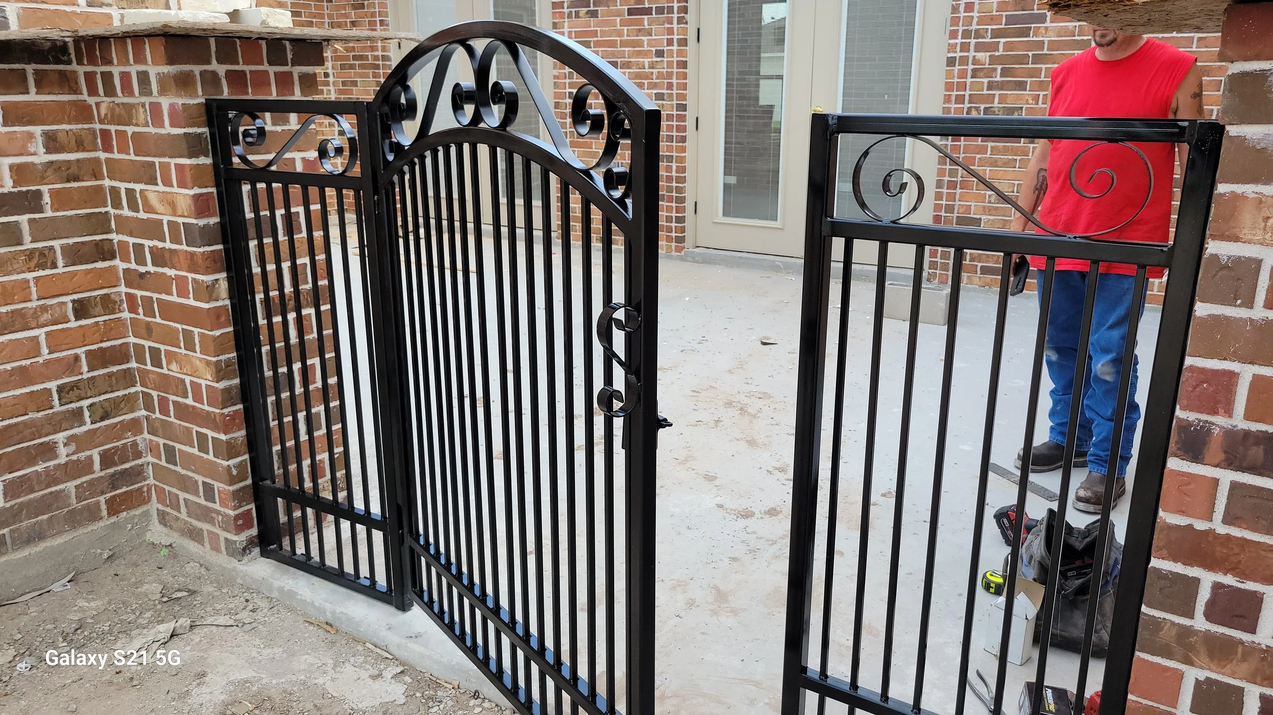 Man in red shirt installing a black metal gate near a brick wall with glass doors in the background.