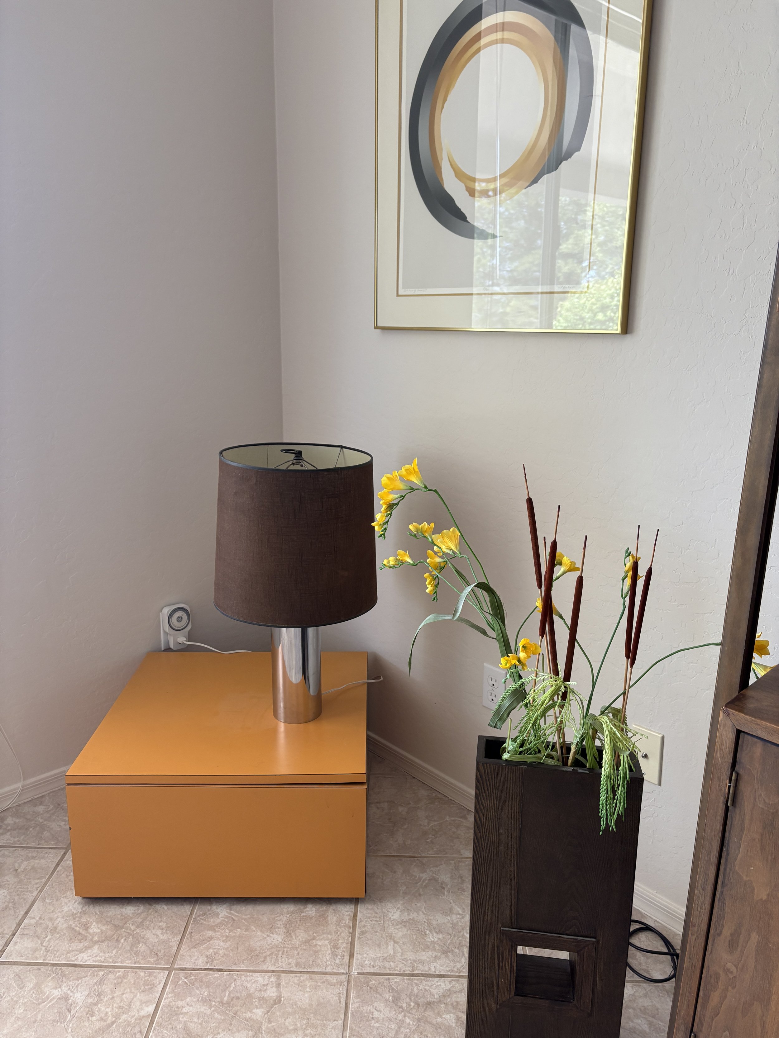 Interior corner with a modern art print, a brown lamp on an orange table, and a tall dark vase with yellow flowers and cattails.