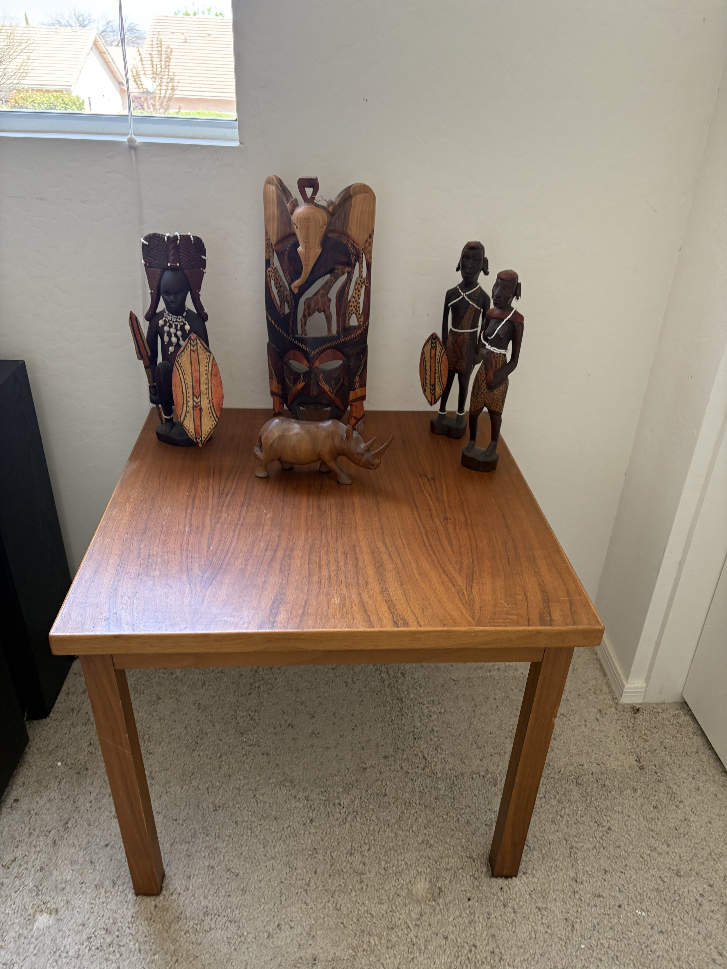 Wooden African-style sculptures on a wooden table, including figures holding shields, a detailed mask, and a rhinoceros carving, against a white wall under a window.