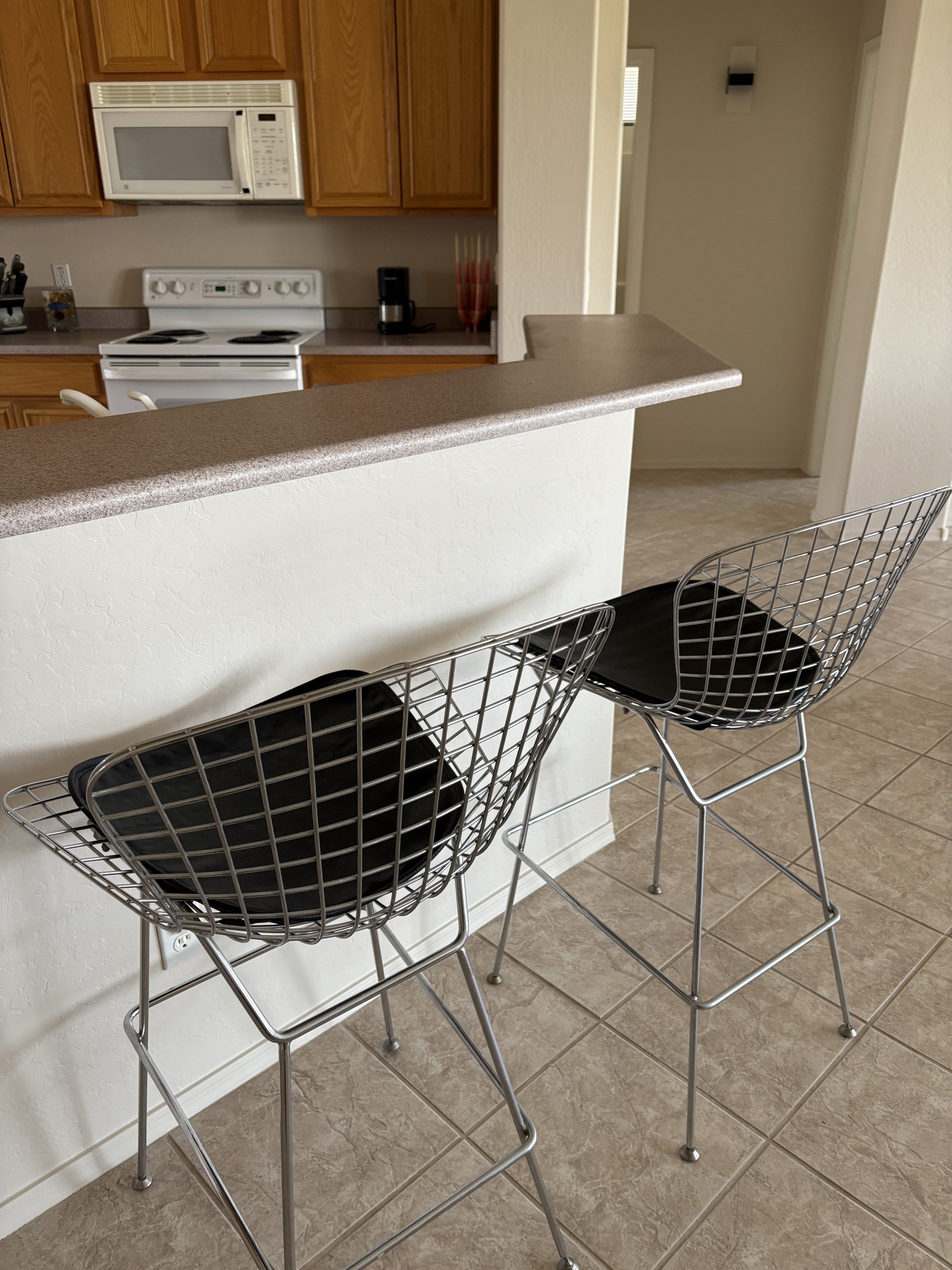 Modern kitchen area with two metal wire barstools, a countertop, and visible appliances including a microwave, stove, and coffee maker.