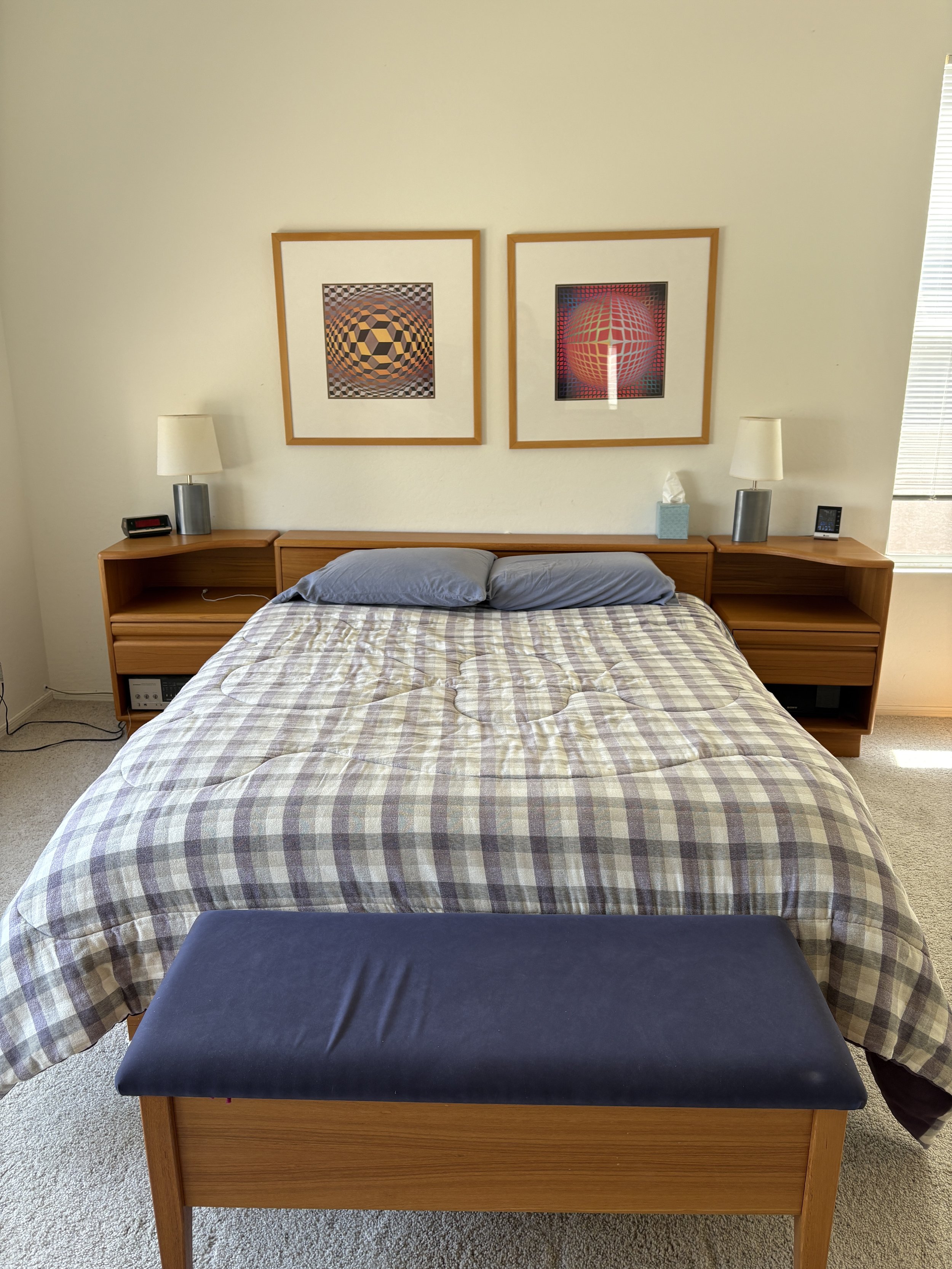 Bedroom with a neatly made bed featuring a plaid comforter and blue pillows, wooden headboard with built-in nightstands, two lamps, and two framed abstract artworks on the wall. A blue cushioned bench is at the foot of the bed.