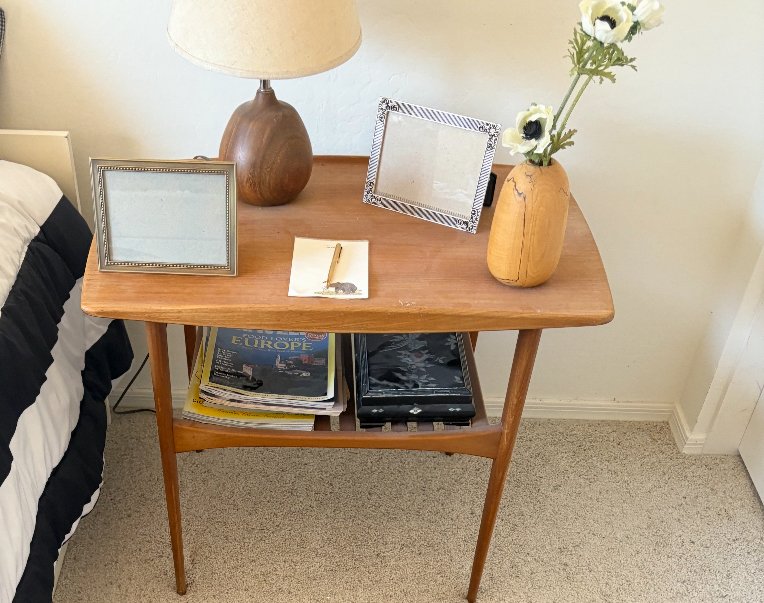 Wooden nightstand with a lamp, empty picture frames, a vase with flowers, a notepad with a pen, magazines, and a book on the lower shelf.