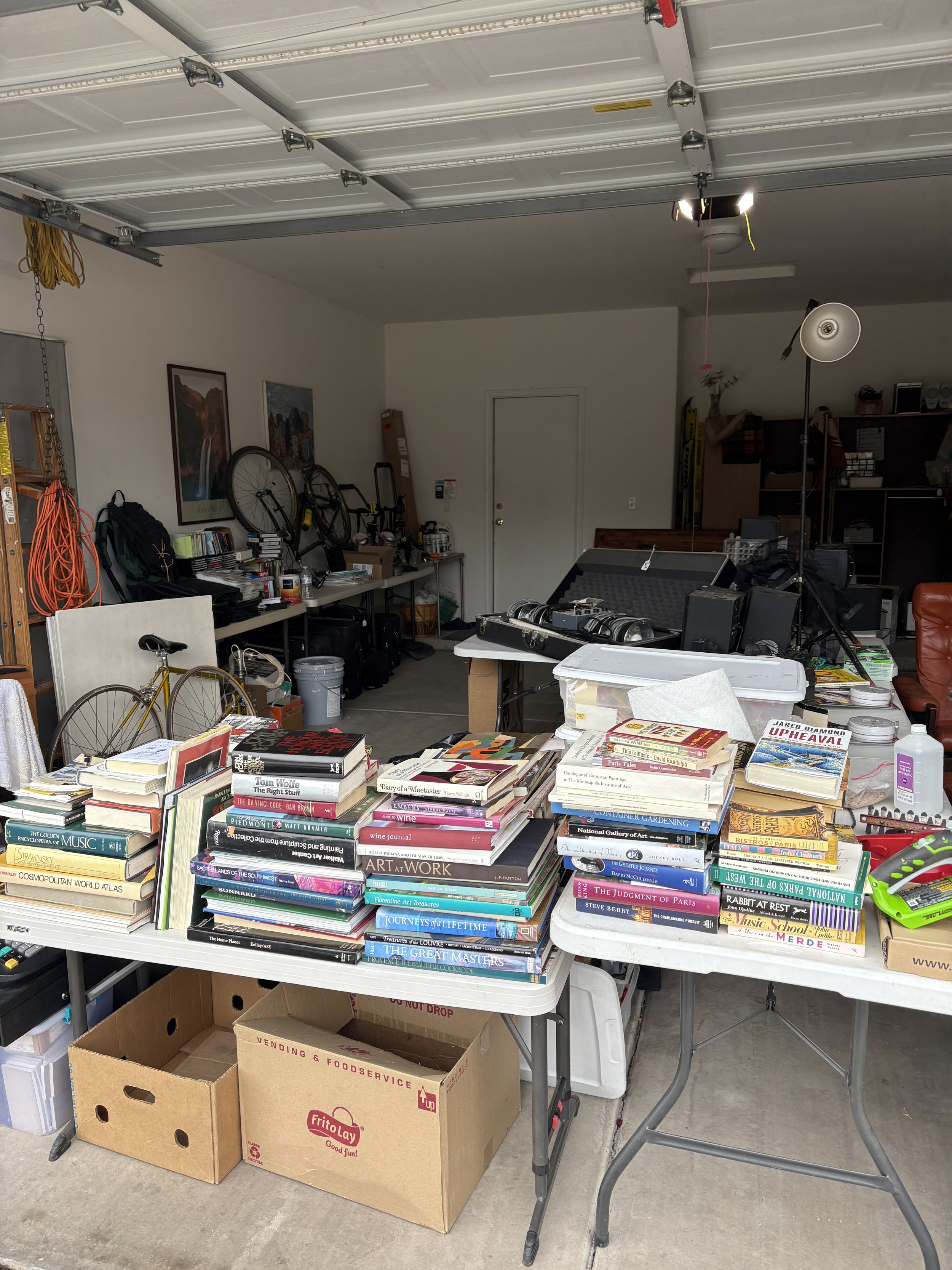 Garage sale setup with tables displaying books, boxes, bicycle, and assorted items in a garage.
