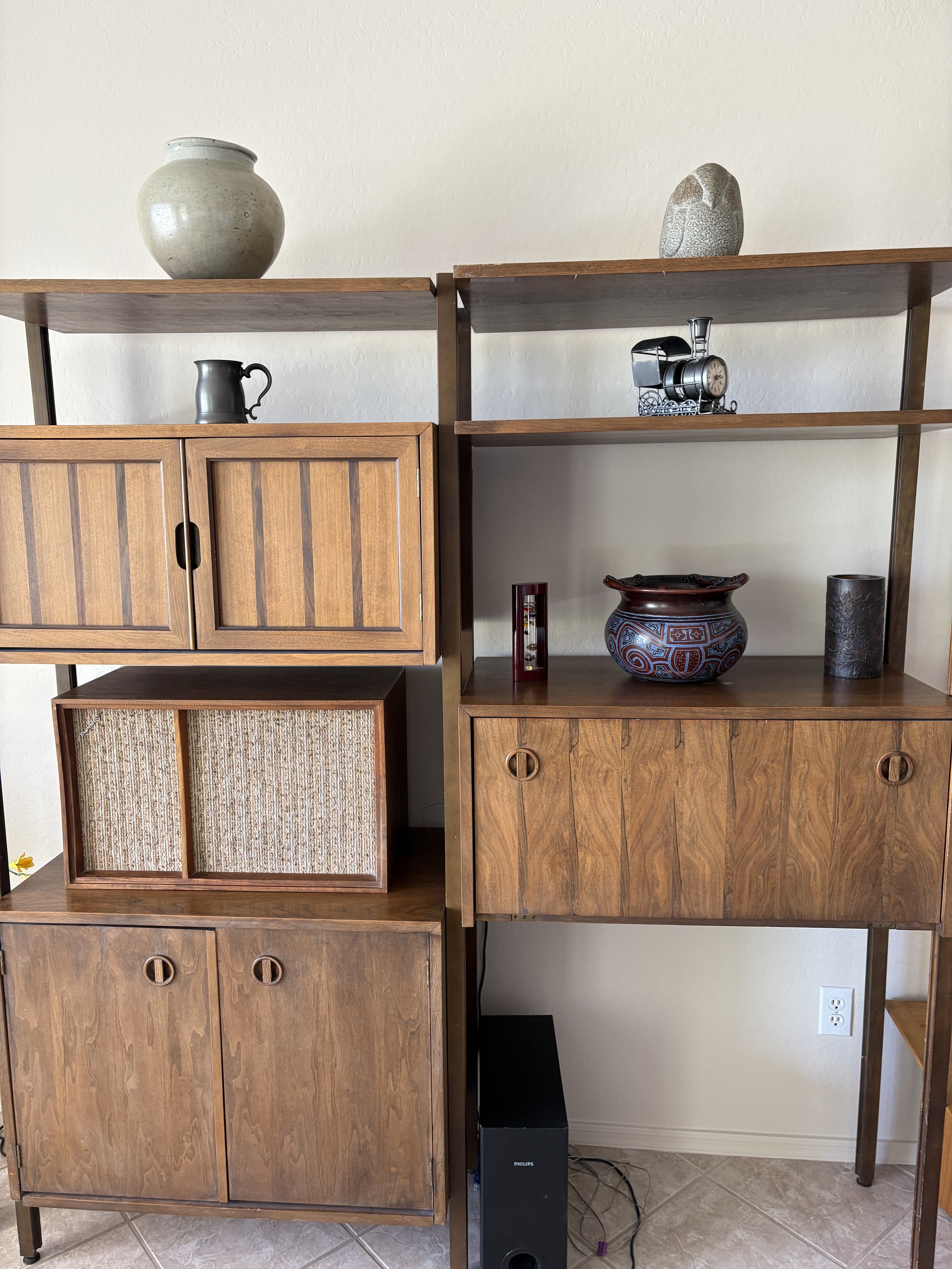 Wooden shelving unit with decorative items including a large vase, small metal mug, train-shaped clock, and patterned ceramic bowl. Bottom cabinets contain speakers and other electronics.