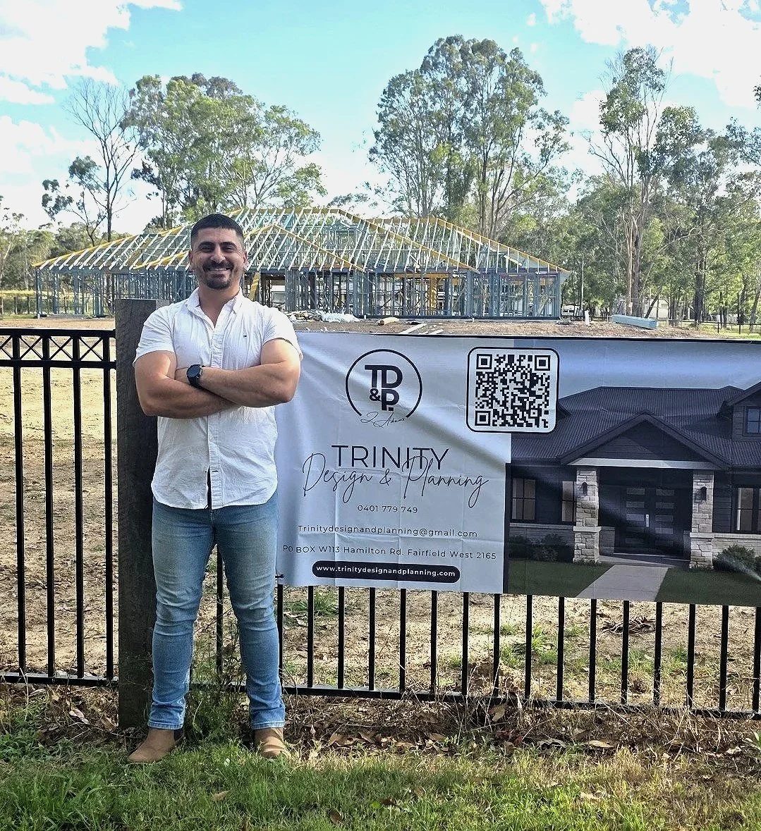 A man standing with crossed arms smiling in front of a sign about Trinity Design and Planning at a construction site with a house illustration and a building frame under construction in the background, among trees and a partly cloudy sky.