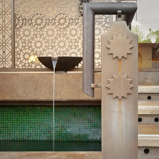 Decorative water fountain with two star-shaped objects, water flowing from a black bowl into a pool with green mosaic tiles, and steps beside it.