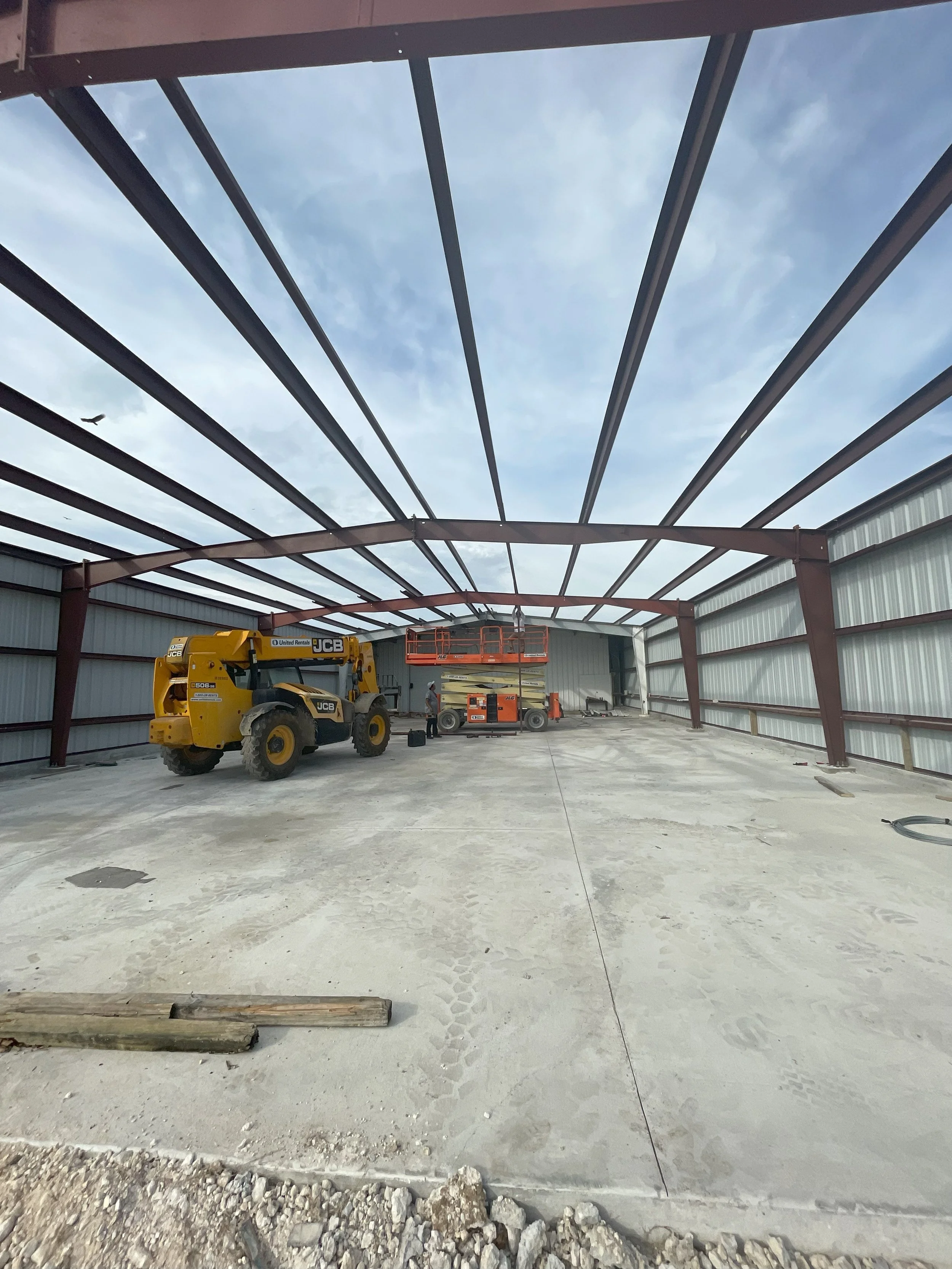 Construction site with steel framework for a building, concrete floor, construction equipment including a yellow JCB loader and an orange scissor lift, and construction worker in the background.